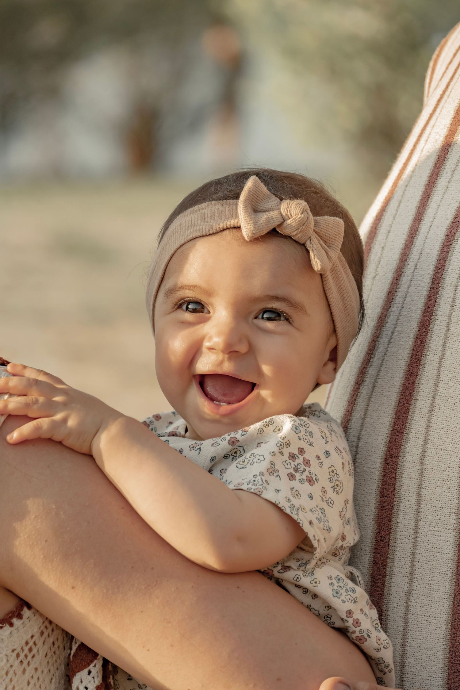 Smiling baby girl wearing a floral outfit and beige bow headband being held outdoors in warm golden light.