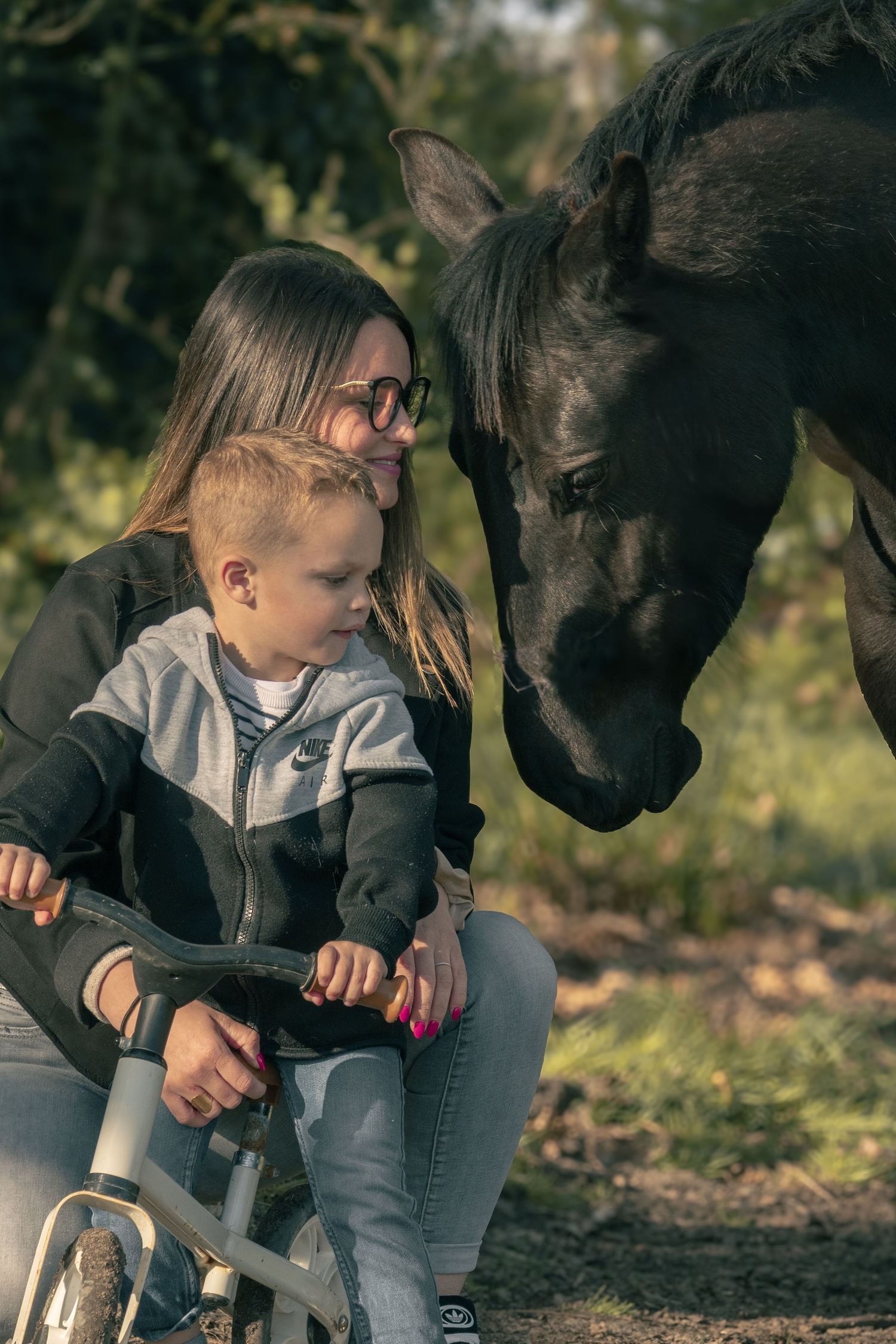 A curious black horse greets a child on a bicycle while accompanied by someone wearing glasses outdoors.