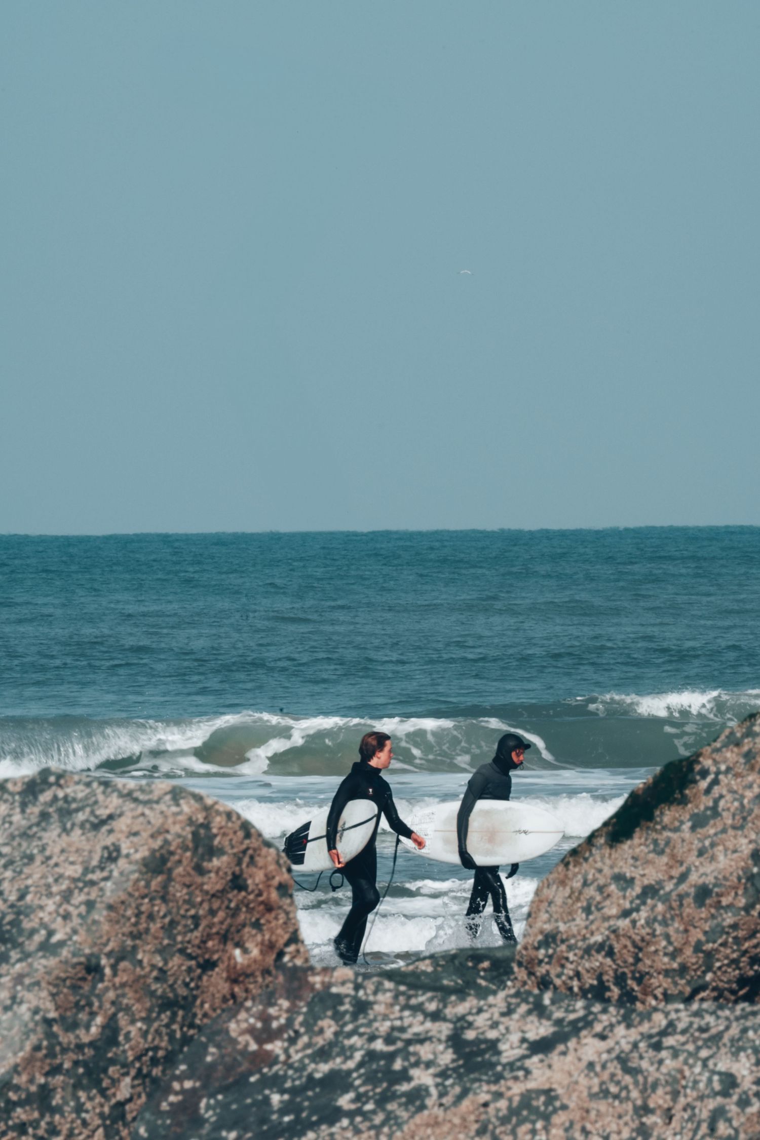 Two surfers standing on rocky coastal outcrop looking out at ocean waves under overcast sky.