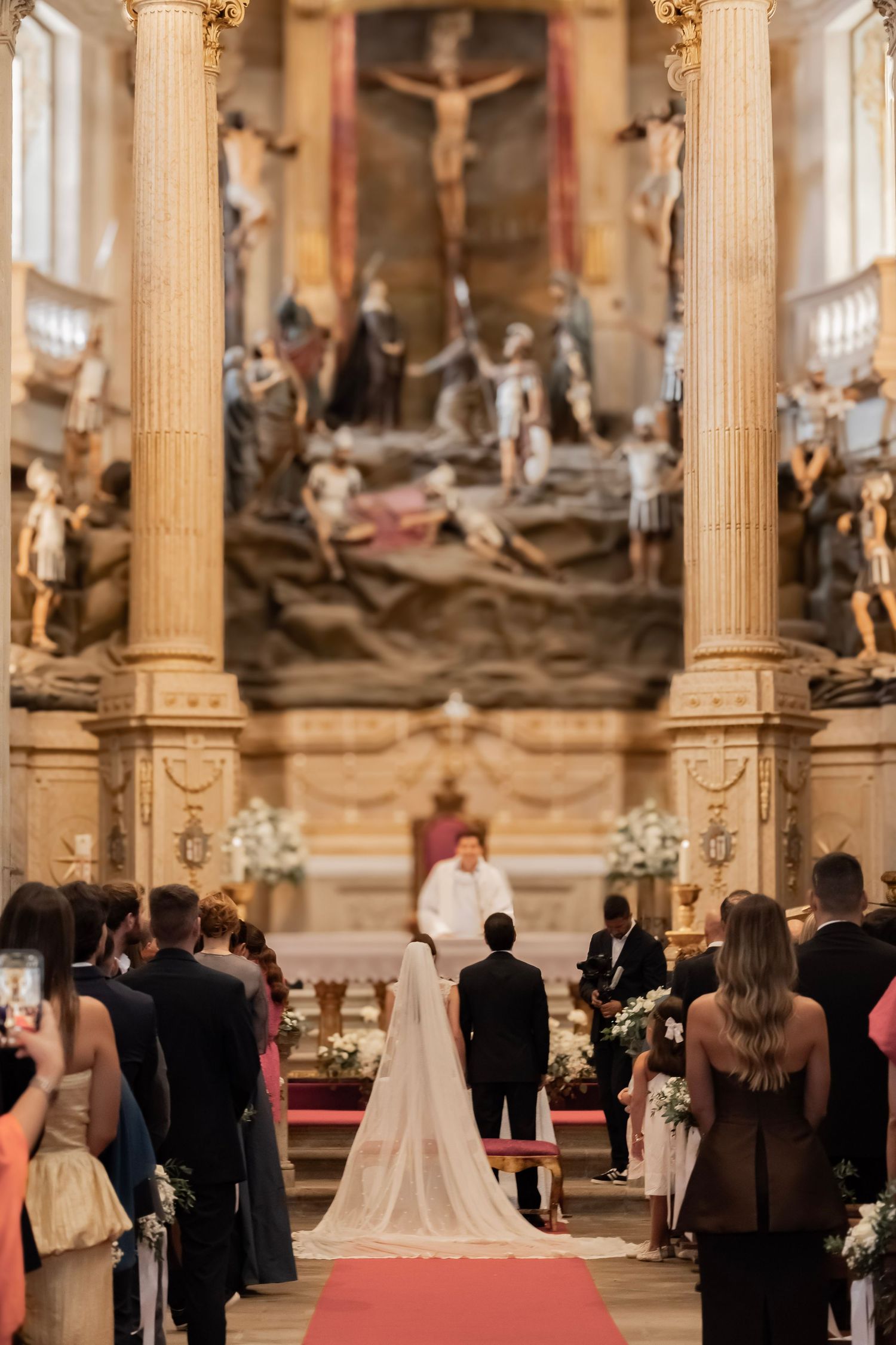 Bride and groom stand at the altar of an ornate church during a wedding ceremony, with guests seated along the aisle.