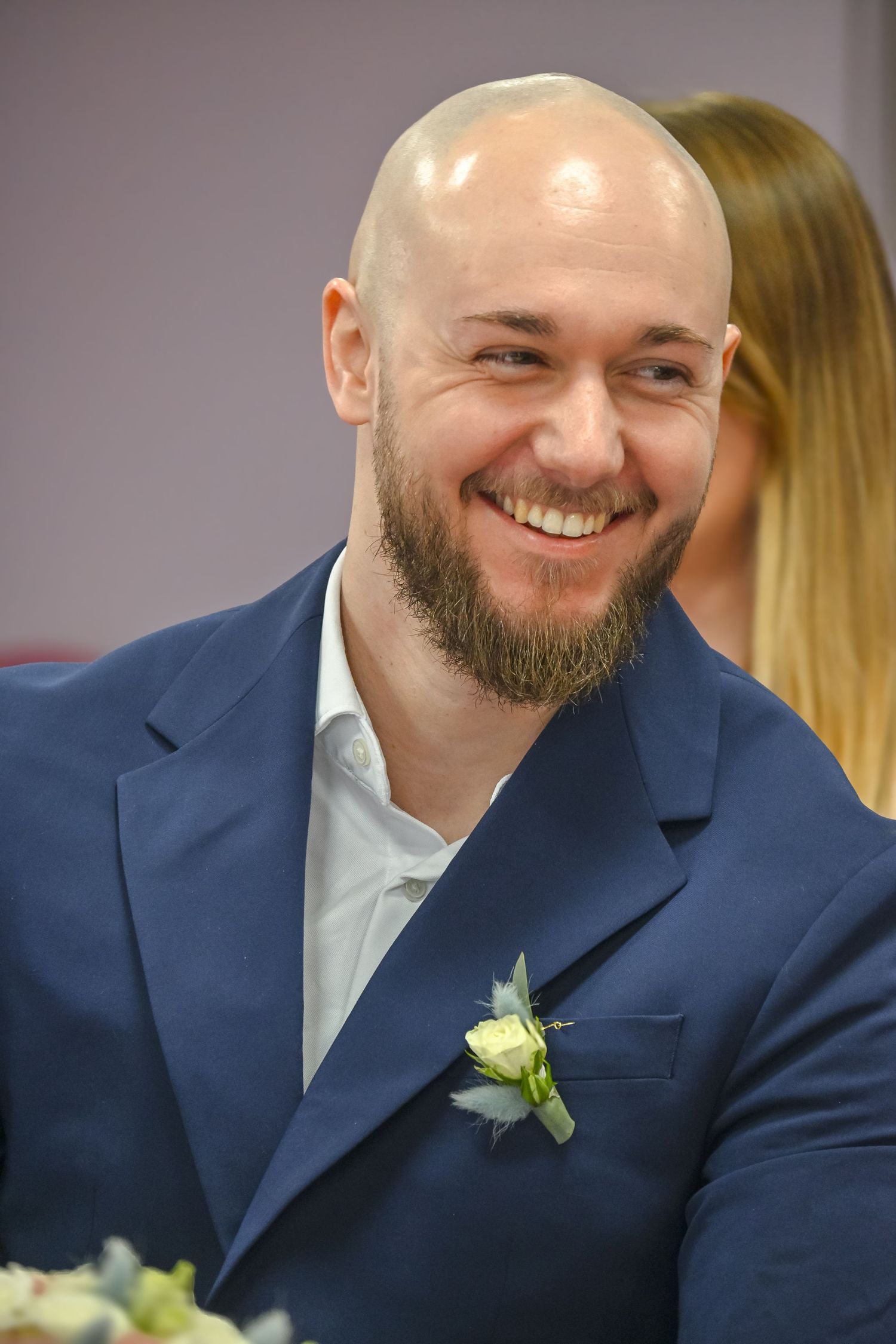 A well-dressed man in navy suit with boutonniere smiles during formal event.