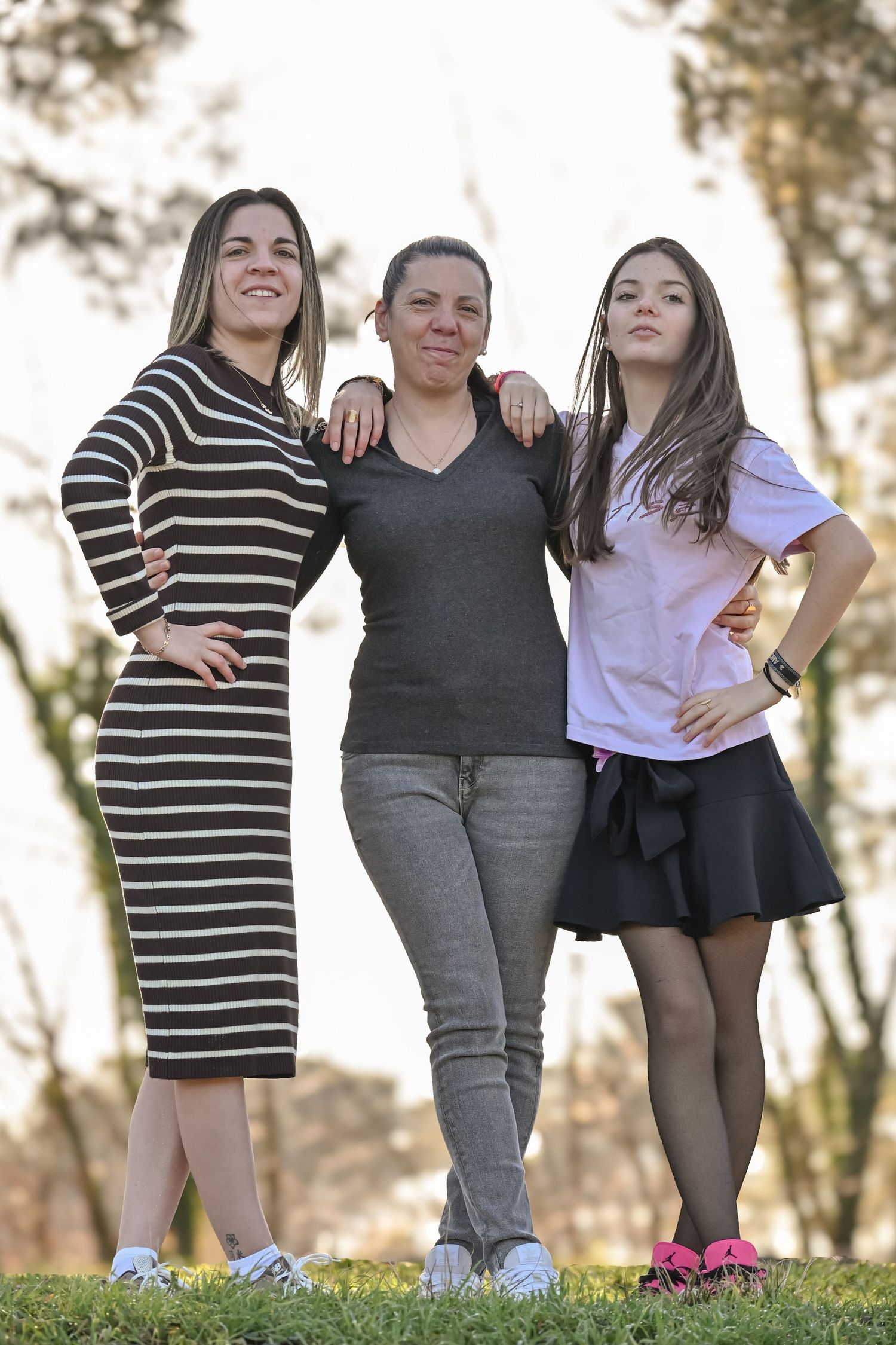 Three friends pose together outdoors in casual attire including a striped dress, gray top, and skirt.