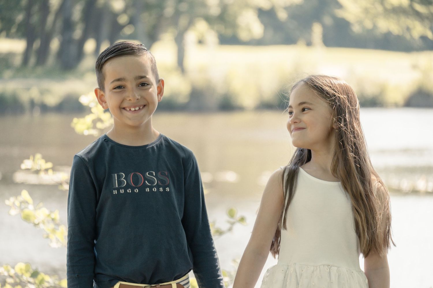 Two children standing together outside on a sunny day by a lake, one wearing a navy sweatshirt and one in white.