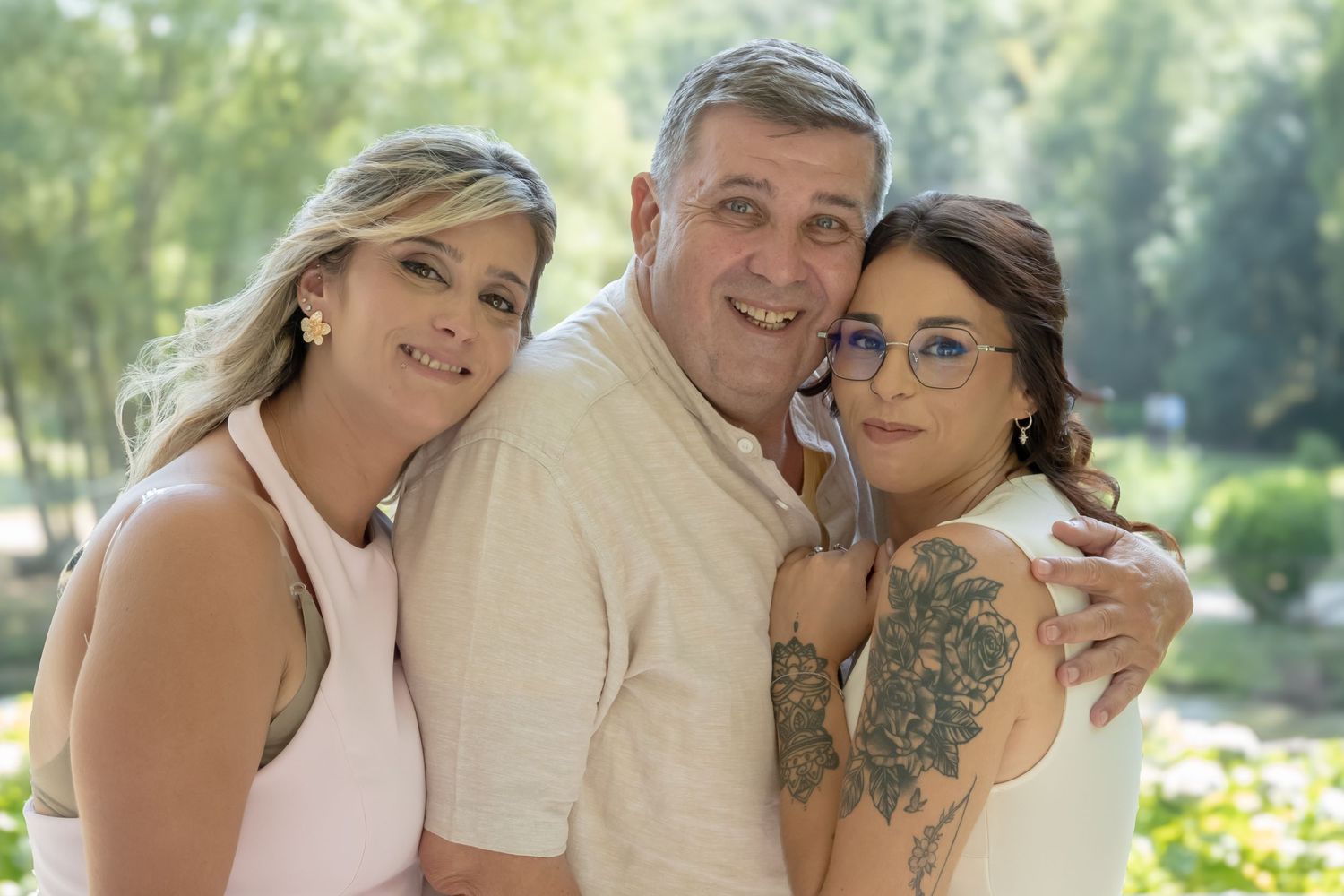 Three people smile together outdoors on a sunny day wearing casual attire in a park setting.