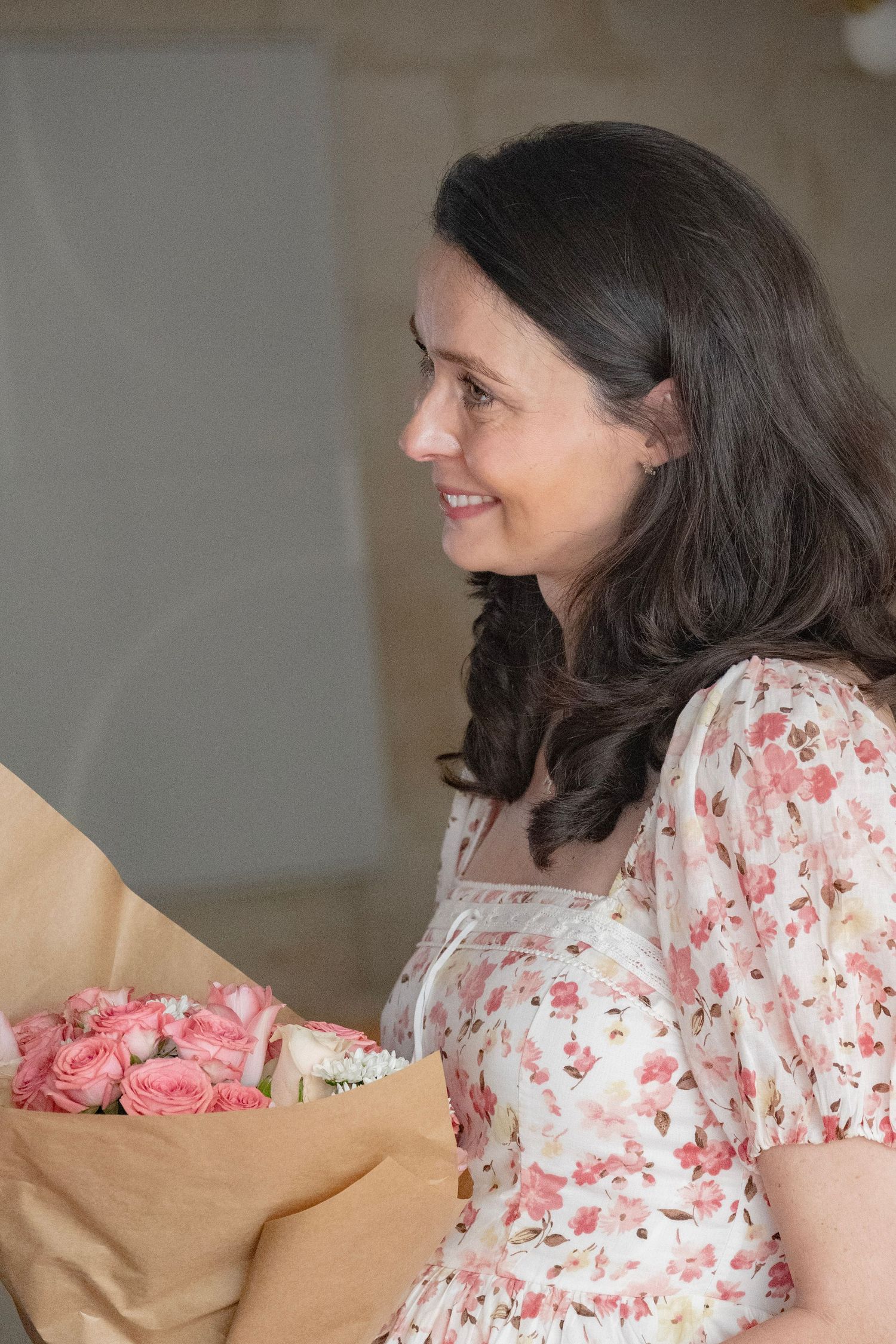 Smiling woman in floral dress holding a bouquet of pink roses wrapped in brown paper.