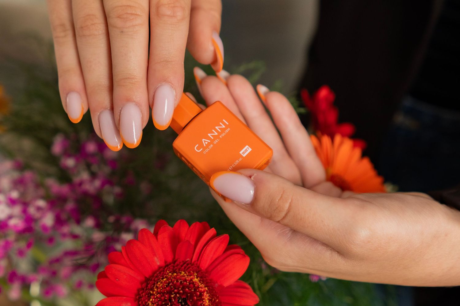 Hands with nude manicure holding an orange CANNI nail product among colorful gerbera daisies.