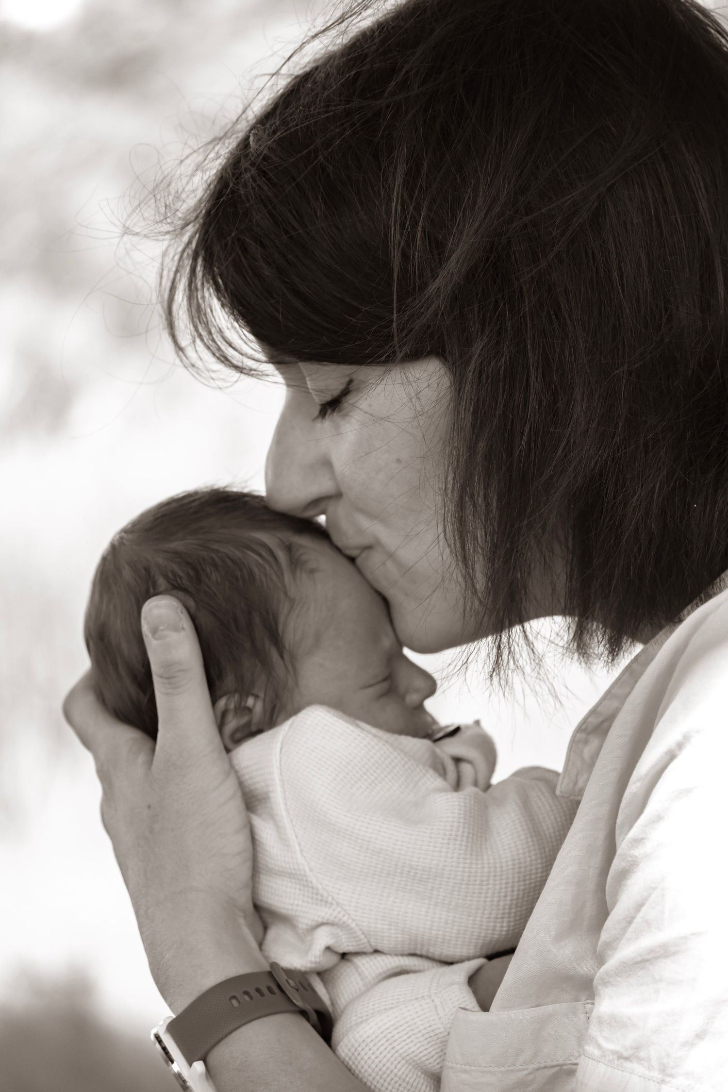 Black and white photo of a mother tenderly kissing her newborn baby on the head while cradling it in her arms.