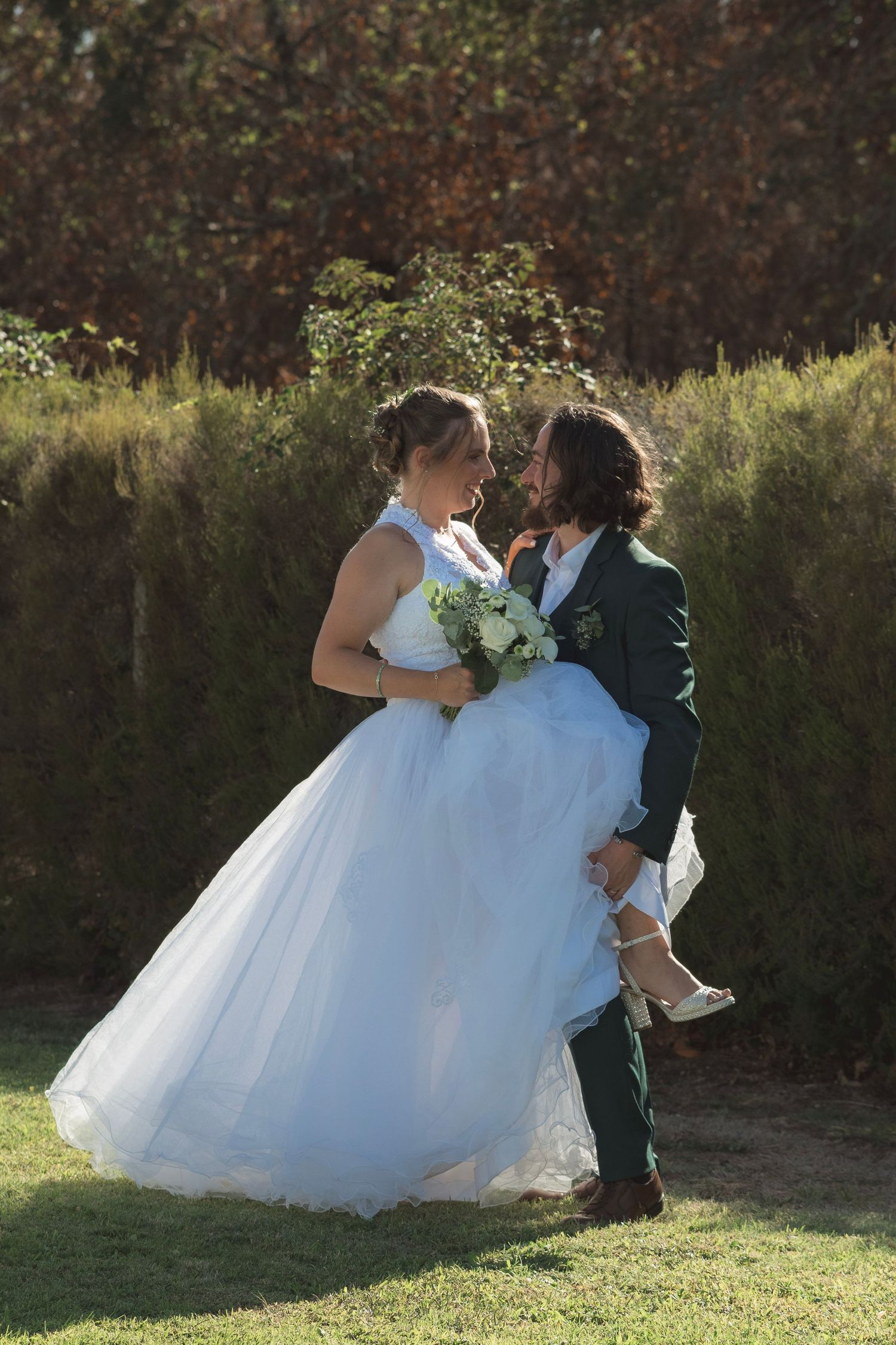 Groom lifting bride in white ballgown and bouquet, sharing a joyful moment outdoors with autumn foliage backdrop.