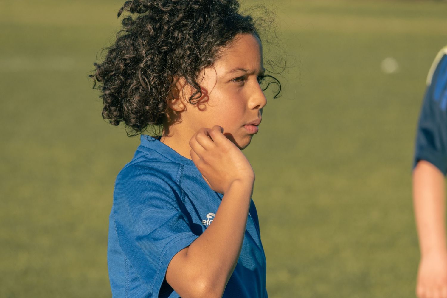 A contemplative athlete in a blue jersey stands pensively with hand on chin during outdoor practice.