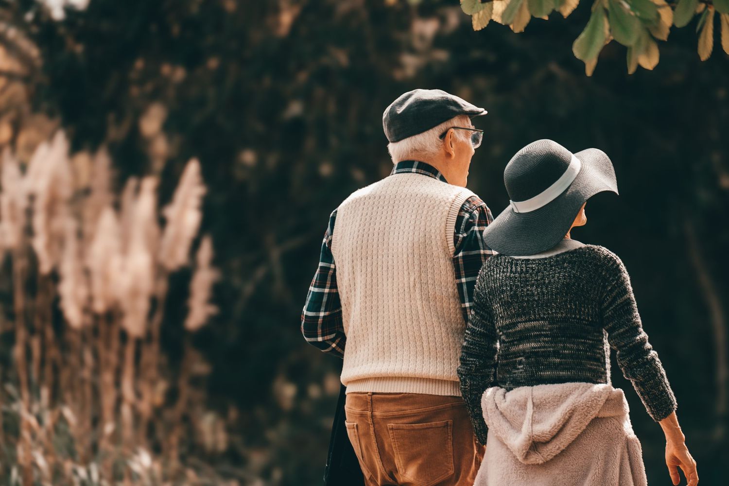 An elderly couple walking together on a path, wearing warm clothing and hats against an autumn backdrop.