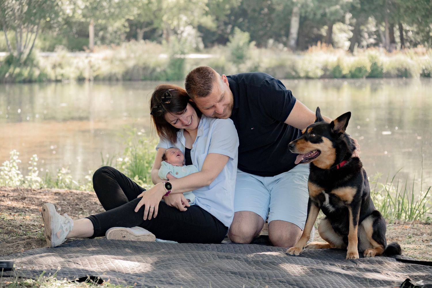 A couple sits by a lake with their newborn baby and a German Shepherd dog during an outdoor family photo session.