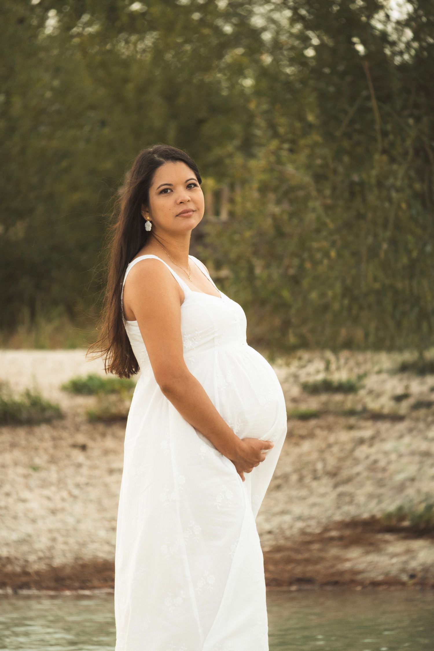 Pregnant woman in white dress holding her baby bump outdoors near water and green trees.