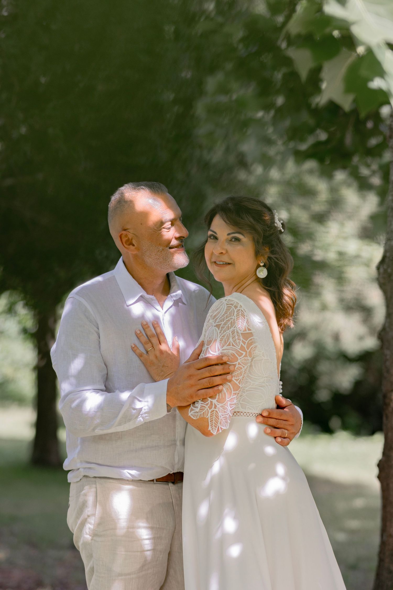 Elegant mature couple embracing outdoors in wedding attire, bride in lace gown and groom in light suit, surrounded by green trees.