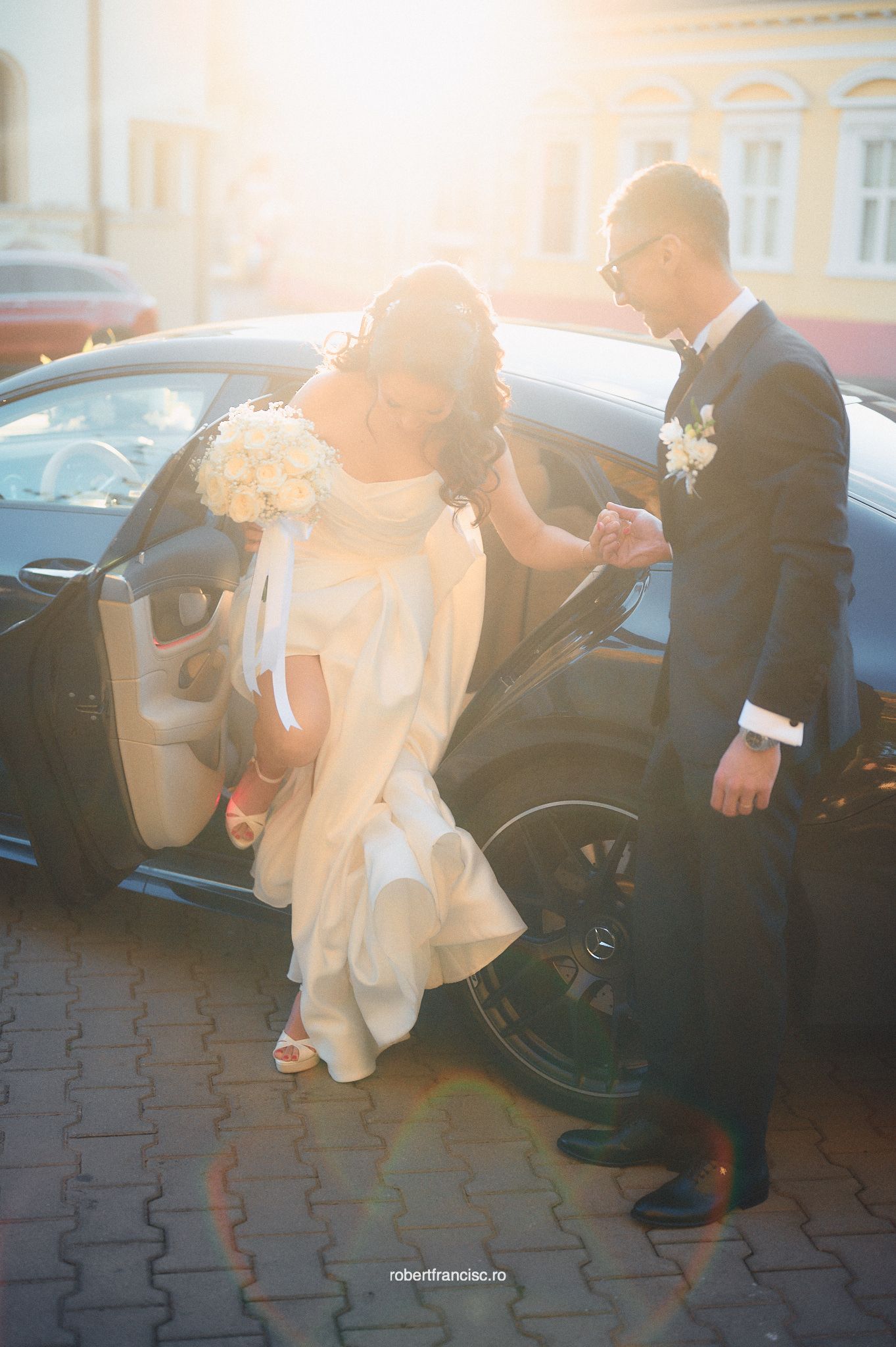 Couple in formal wedding attire shares a tender moment in sunlit parking lot with lens flare effect.