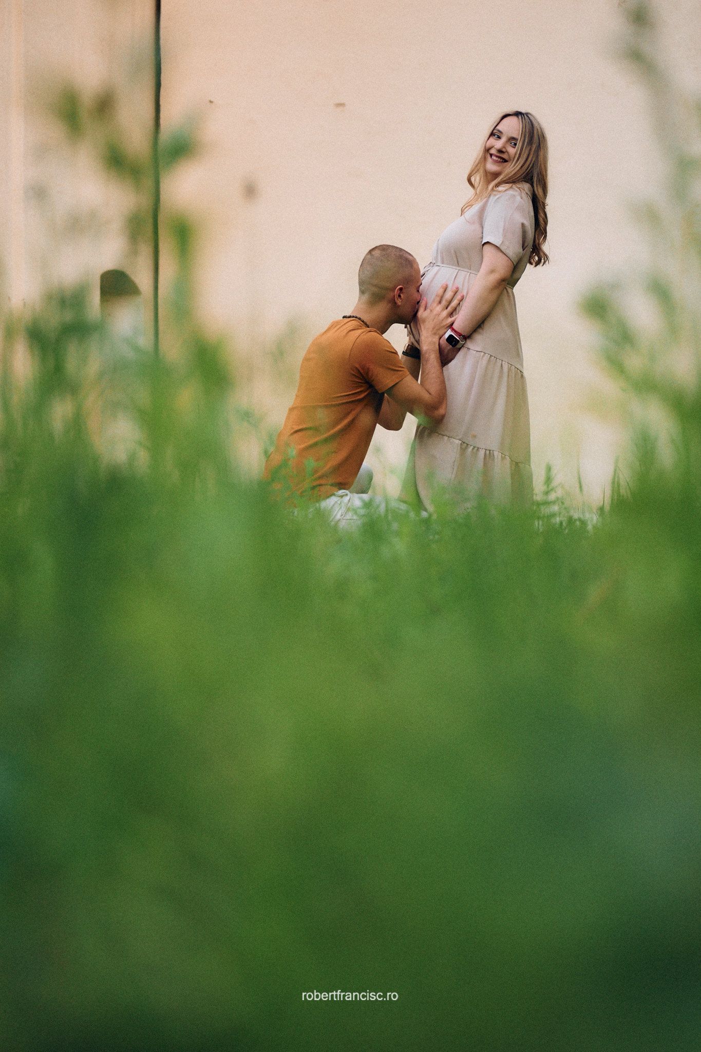 A tender maternity photo captured through green grass showing an expectant couple sharing an intimate moment outdoors.