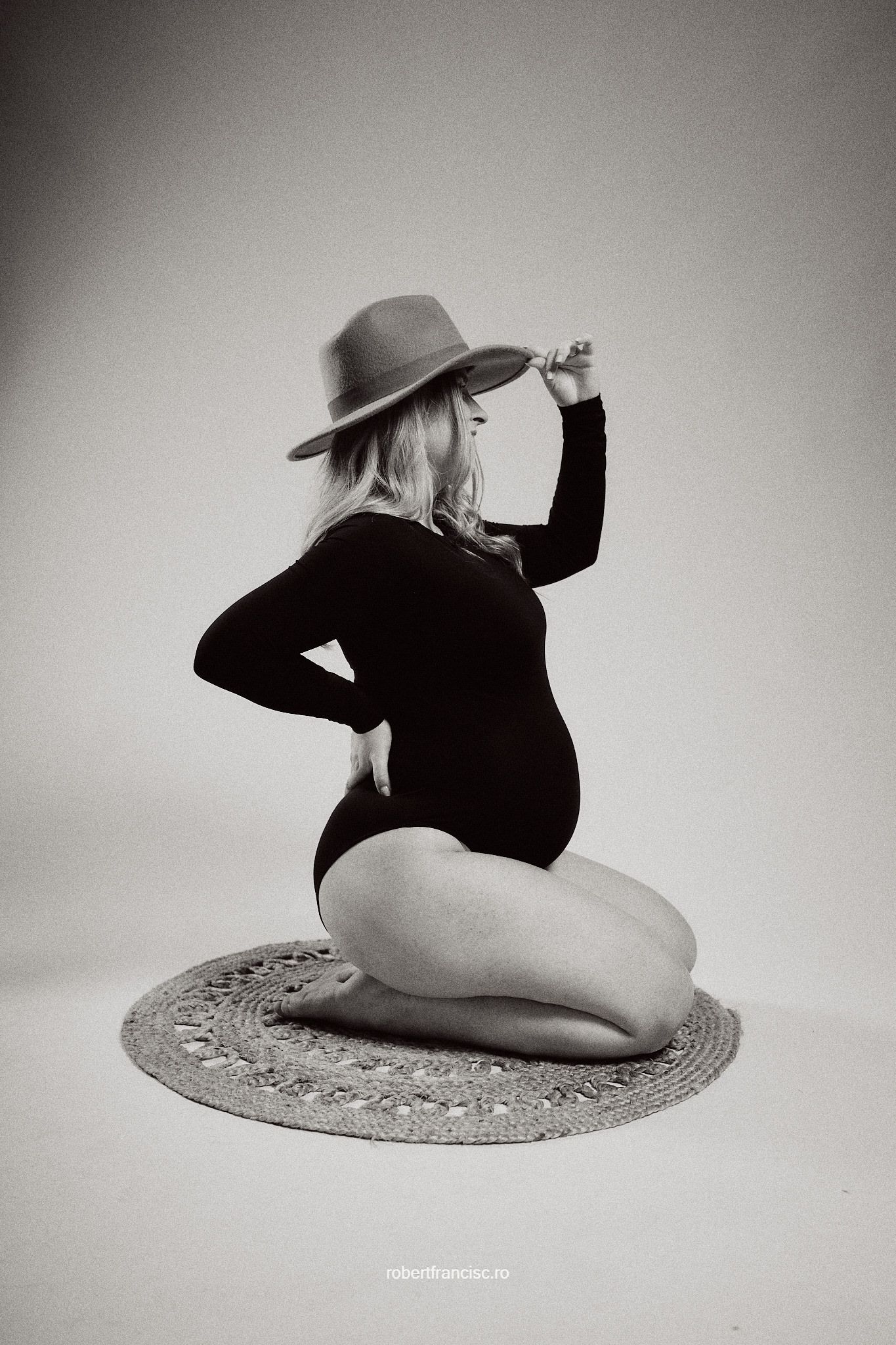Black and white artistic maternity photo of a woman in a black bodysuit and hat posing on a decorative rug.