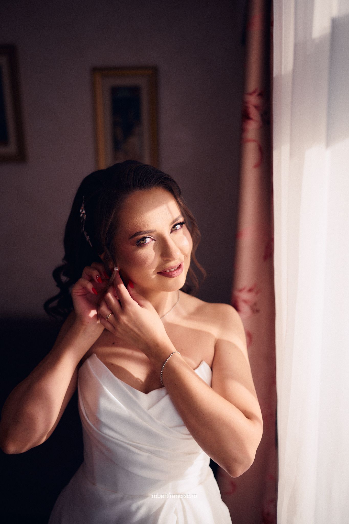 A bride in a white strapless wedding dress stands by a window in soft natural light, adjusting her earring.
