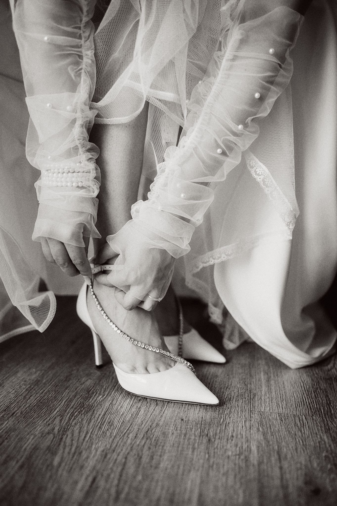 Close-up black and white shot of hands fastening elegant white bridal shoes with delicate tulle fabric details.