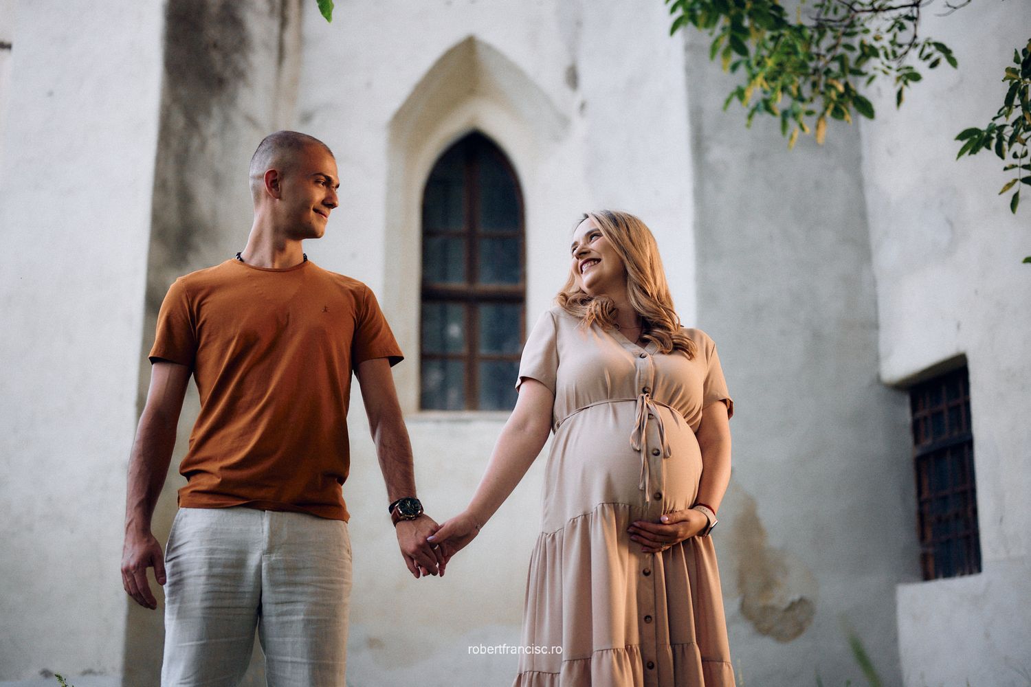 Couple in casual attire walks hand in hand along a white stone wall with arched windows and green foliage above.