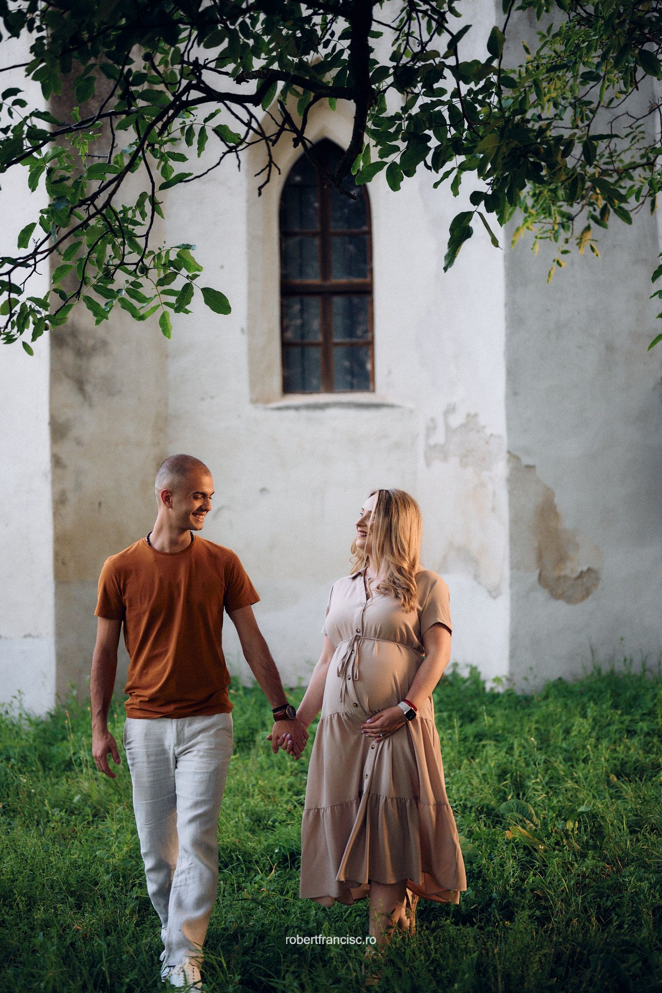 A couple standing together near a white church wall with arched window and green foliage overhead.