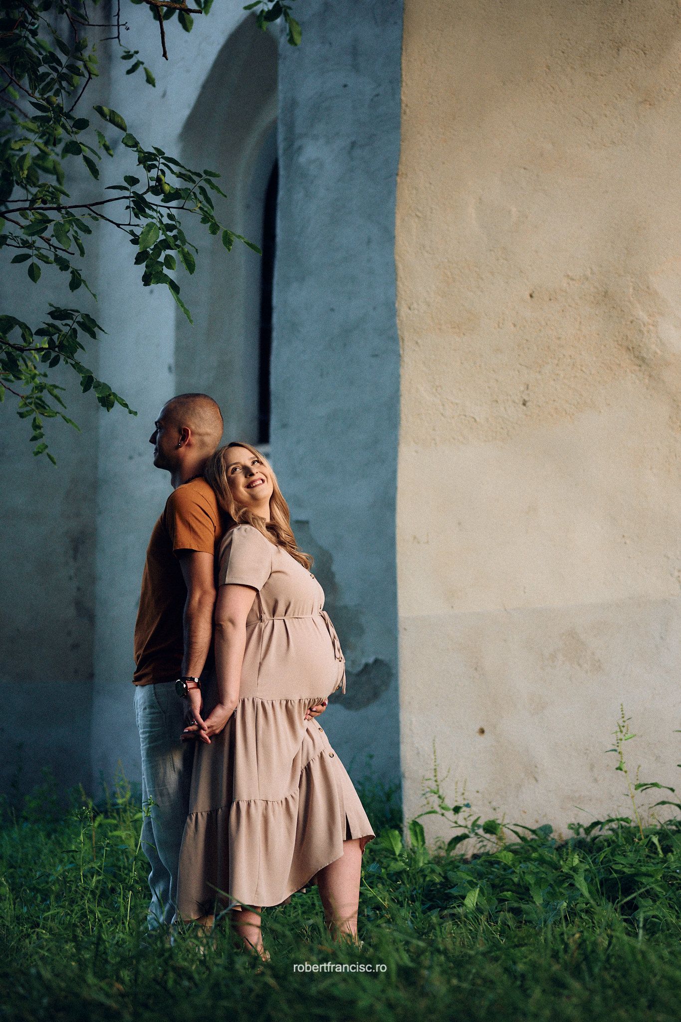 A couple embraces romantically against a blue wall with green grass in a series of sweet outdoor portraits.