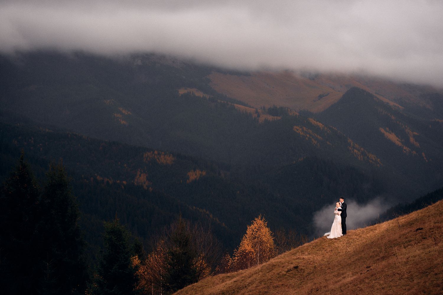 A distant wedding couple silhouetted against misty mountain peaks at sunset.