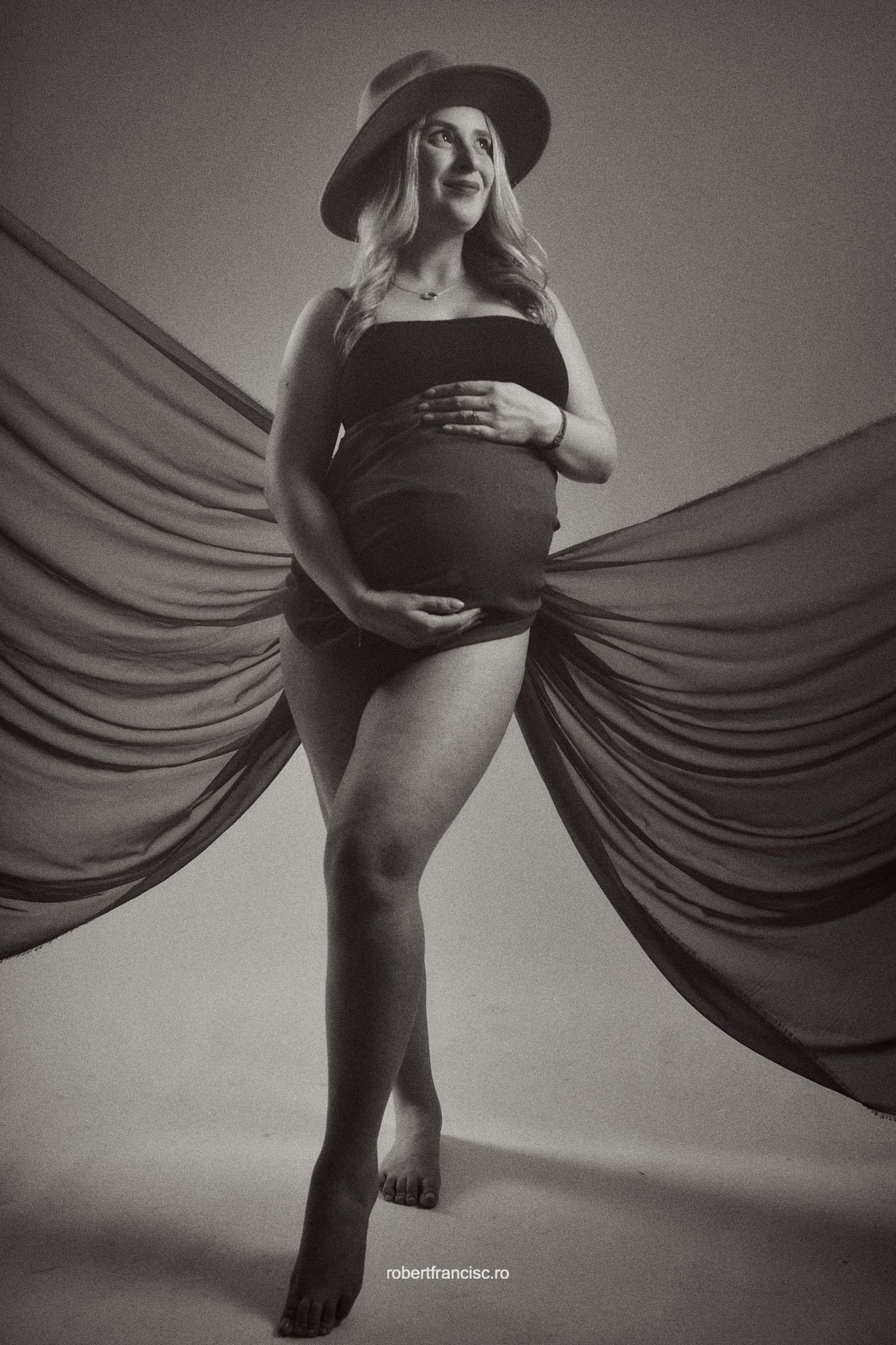 Artistic black and white maternity photo with flowing fabric and hat creating dramatic silhouette against light background.