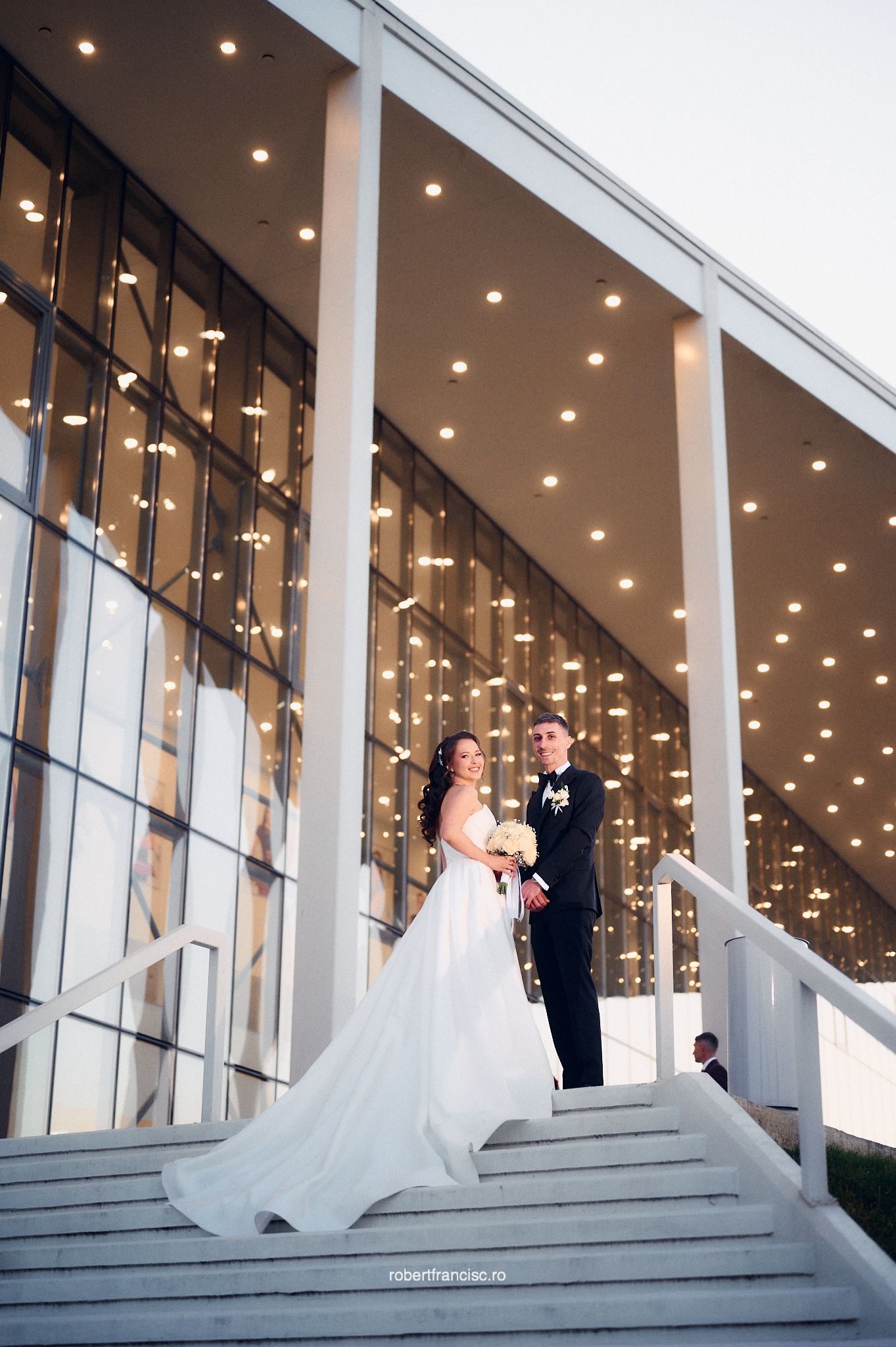 A couple in formal wedding attire poses on modern architectural steps with illuminated ceiling lights in the background.
