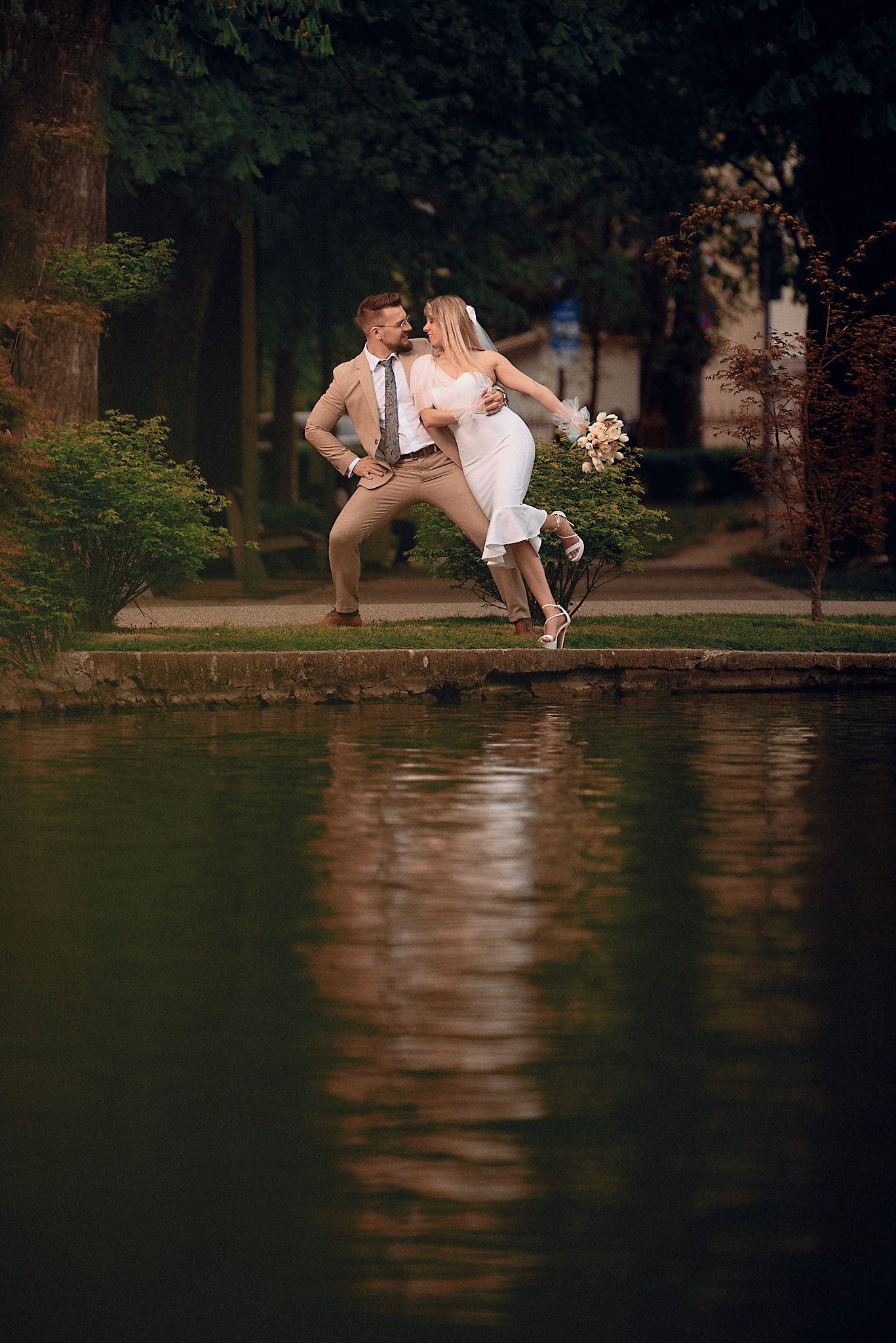 Playful wedding couple jumping by pond in park with water reflection.