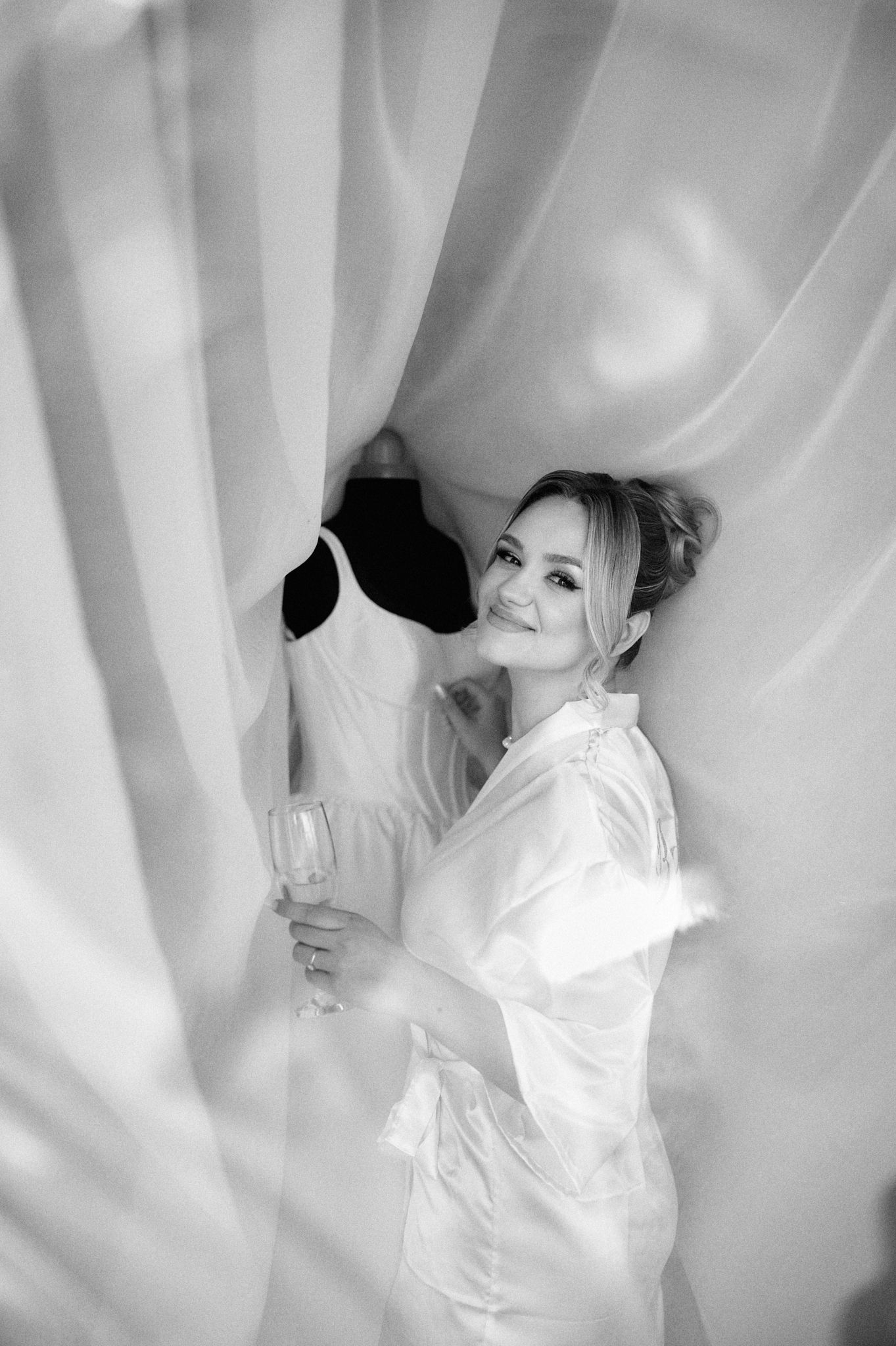 Black and white photo of person in white robe holding champagne glass next to wedding dress.