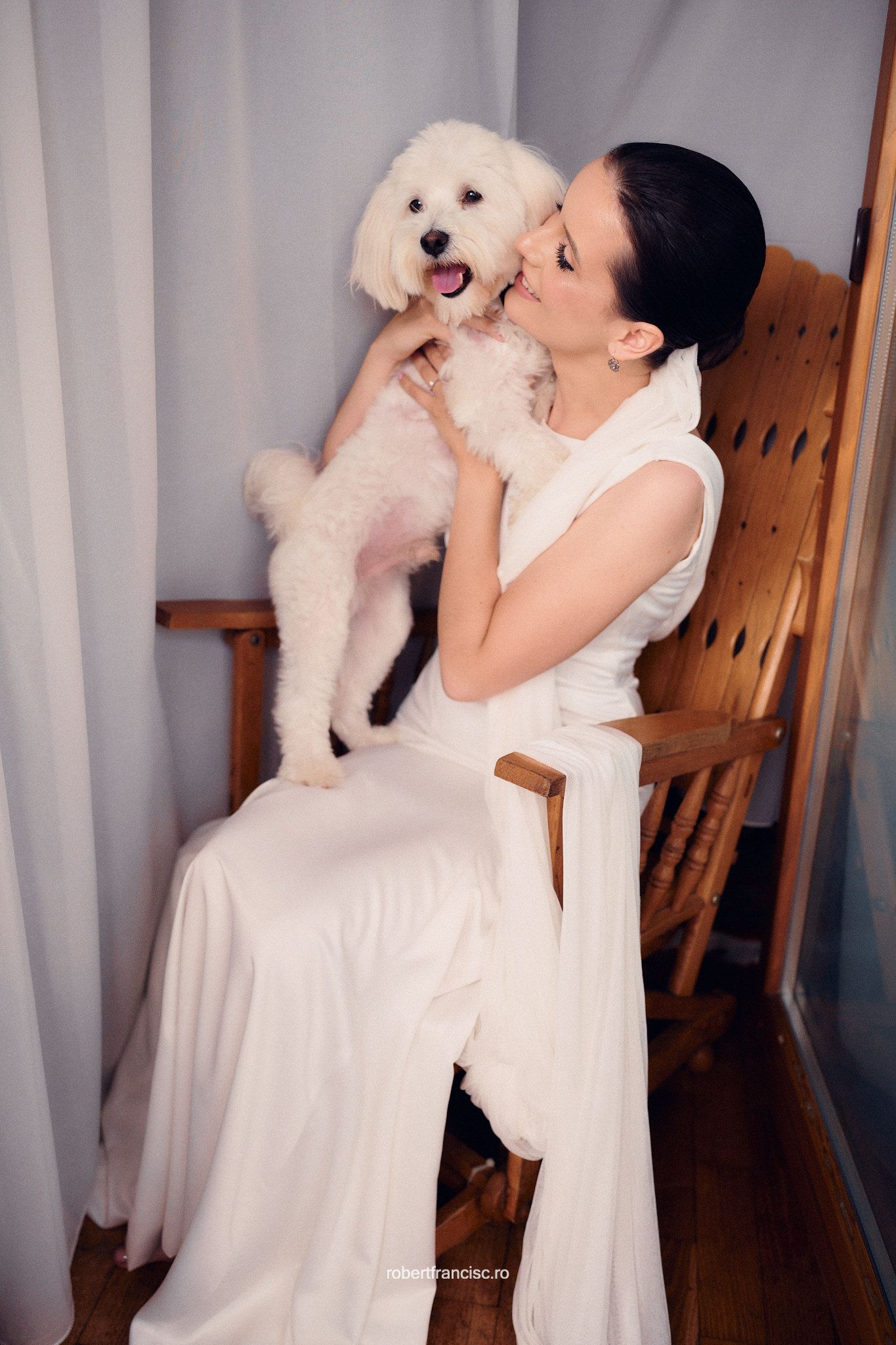 A white fluffy dog shares an affectionate moment with someone in a white dress while sitting in a wooden rocking chair.