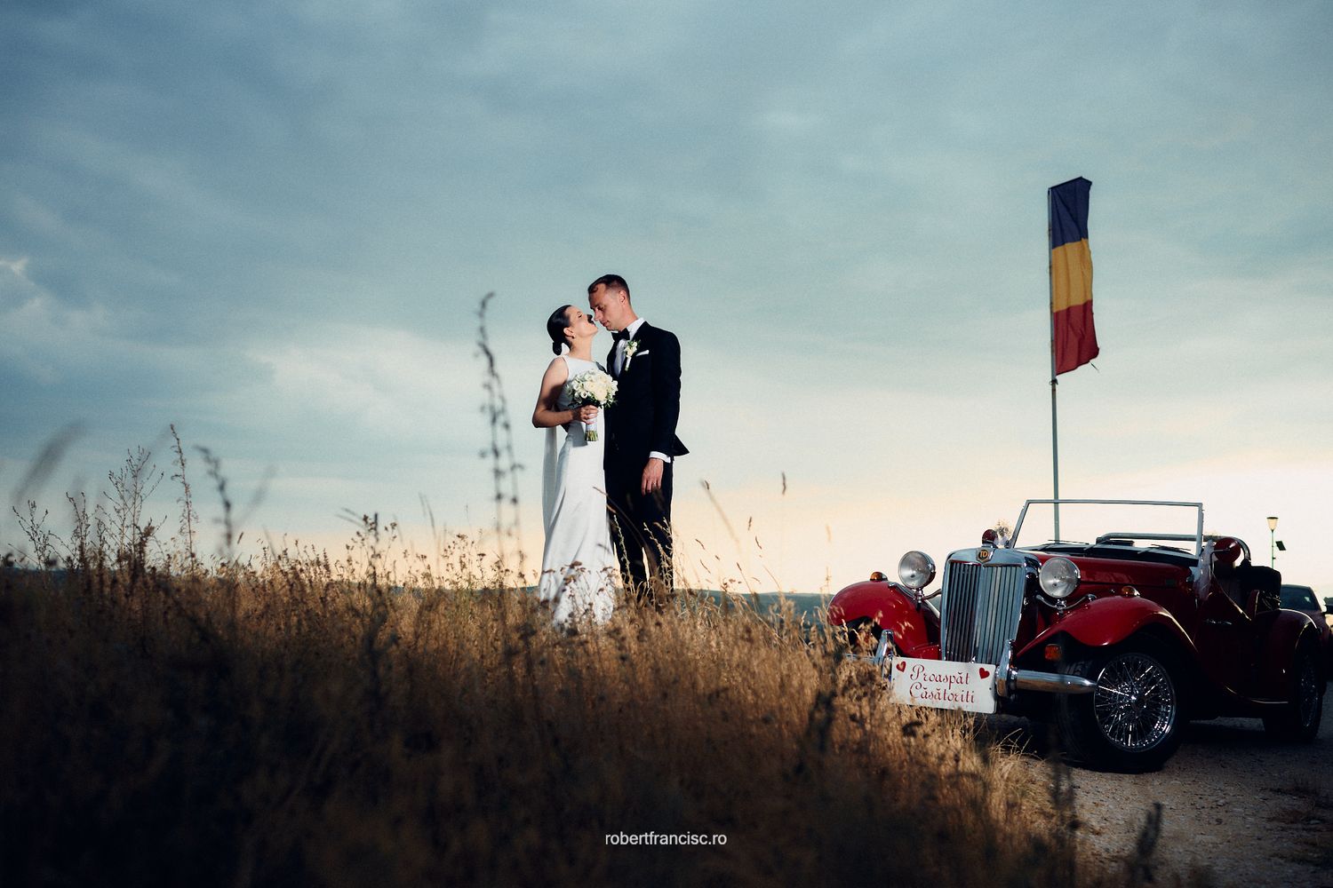 Vintage car parked on a hilltop at sunset with a couple and colorful flag visible in the scene.