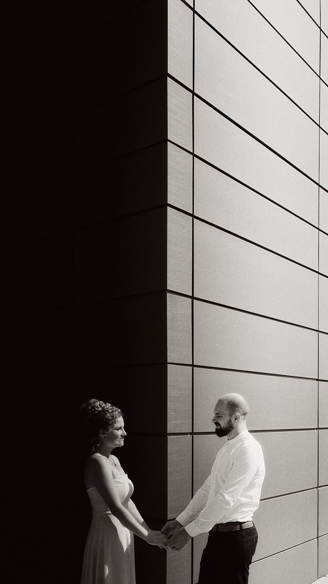 Artistic black and white architectural photo of couple holding hands against modern building wall.
