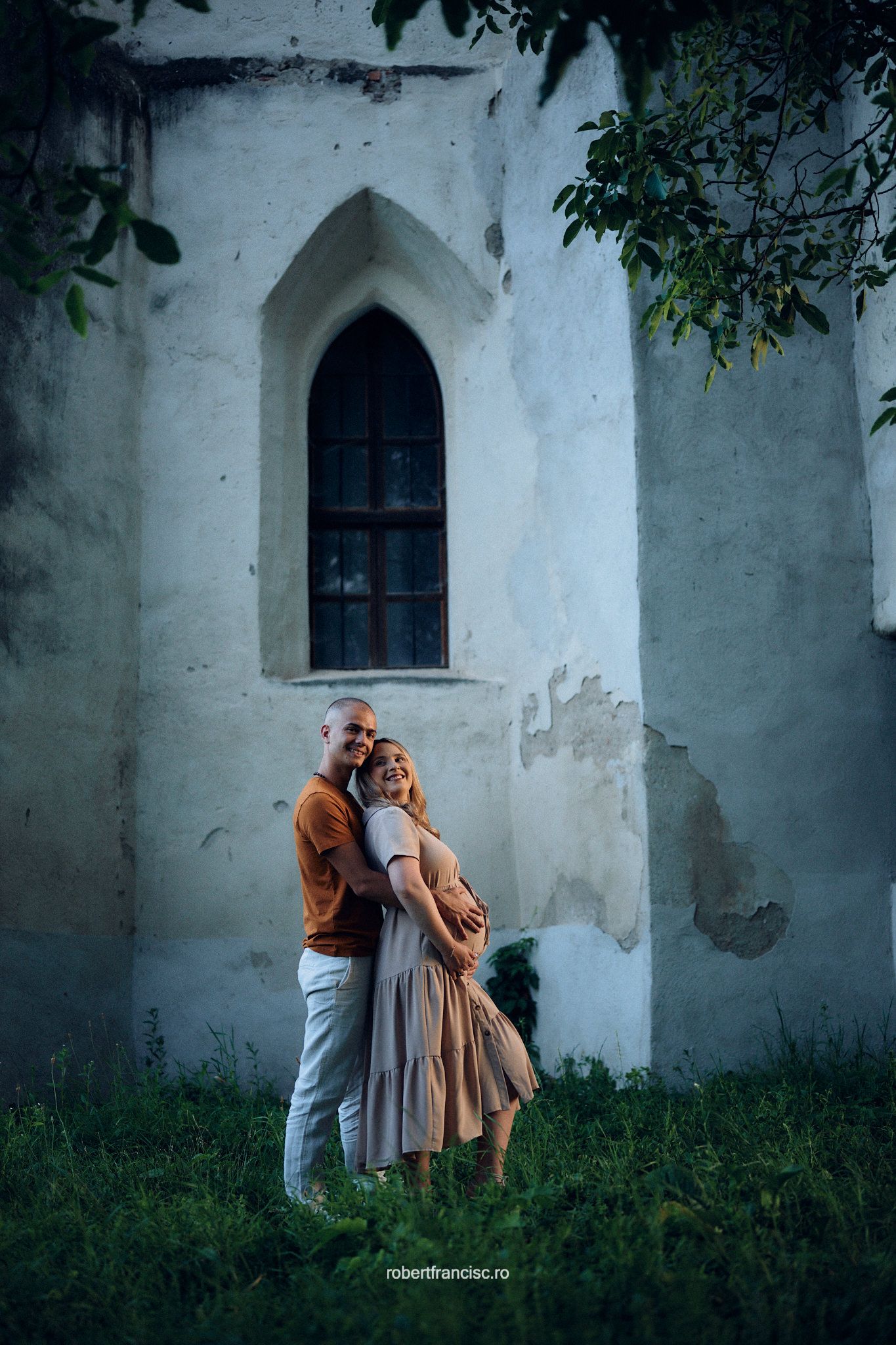 A romantic couple embraces against a white church wall with Gothic arched window and green grass on a moody evening.