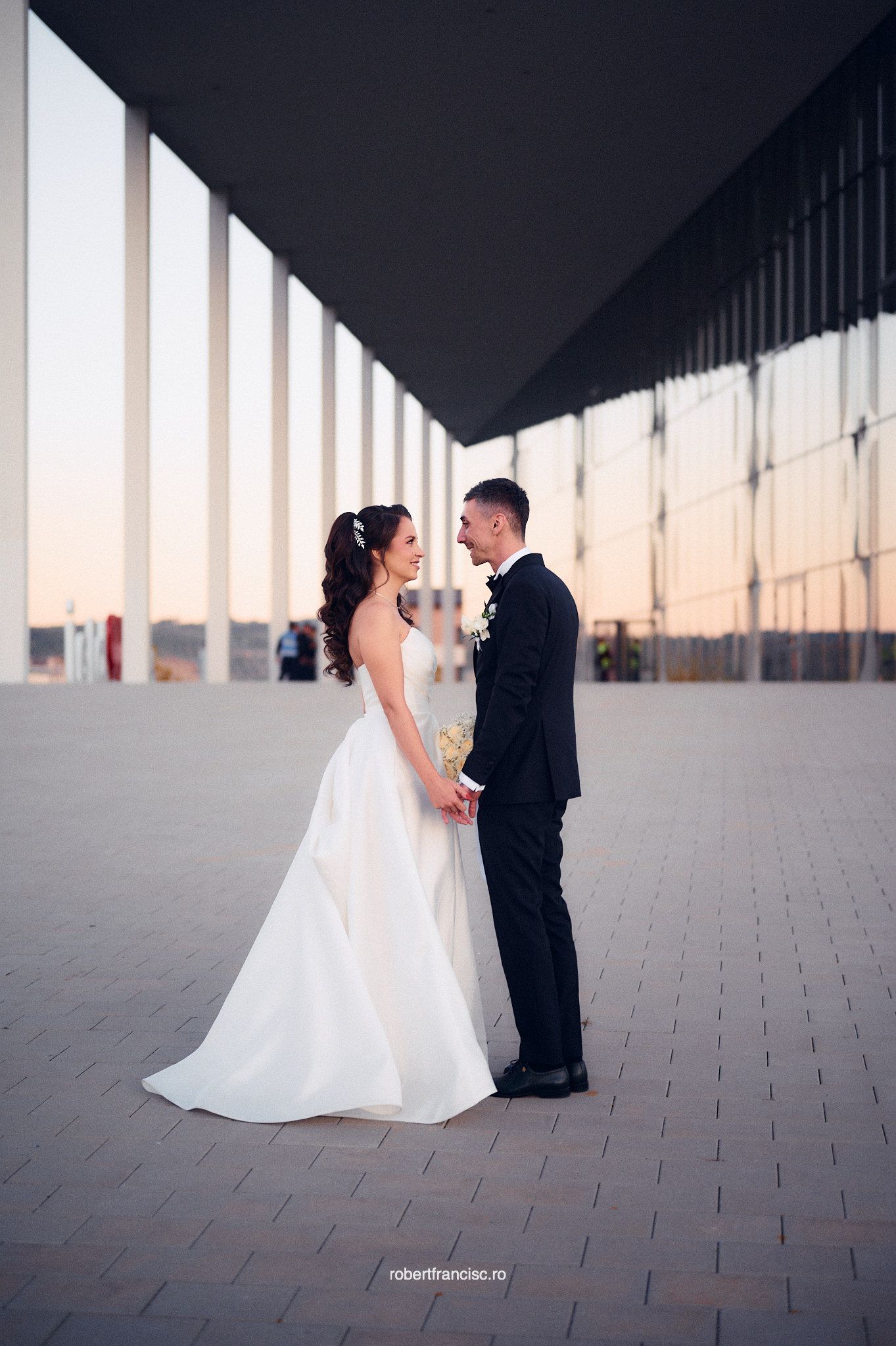 A couple in wedding attire stands together under a modern architectural overhang at sunset.