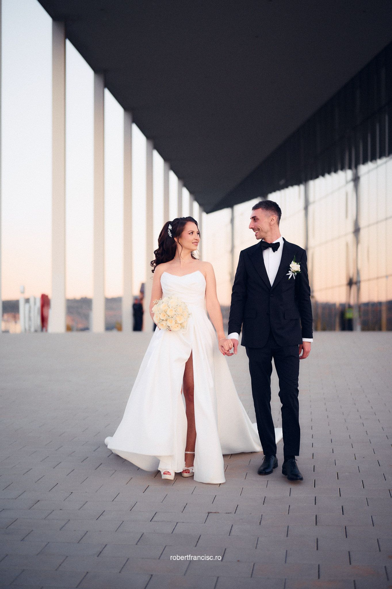 A bride in white and groom in black suit walk together through a modern architectural corridor at sunset.