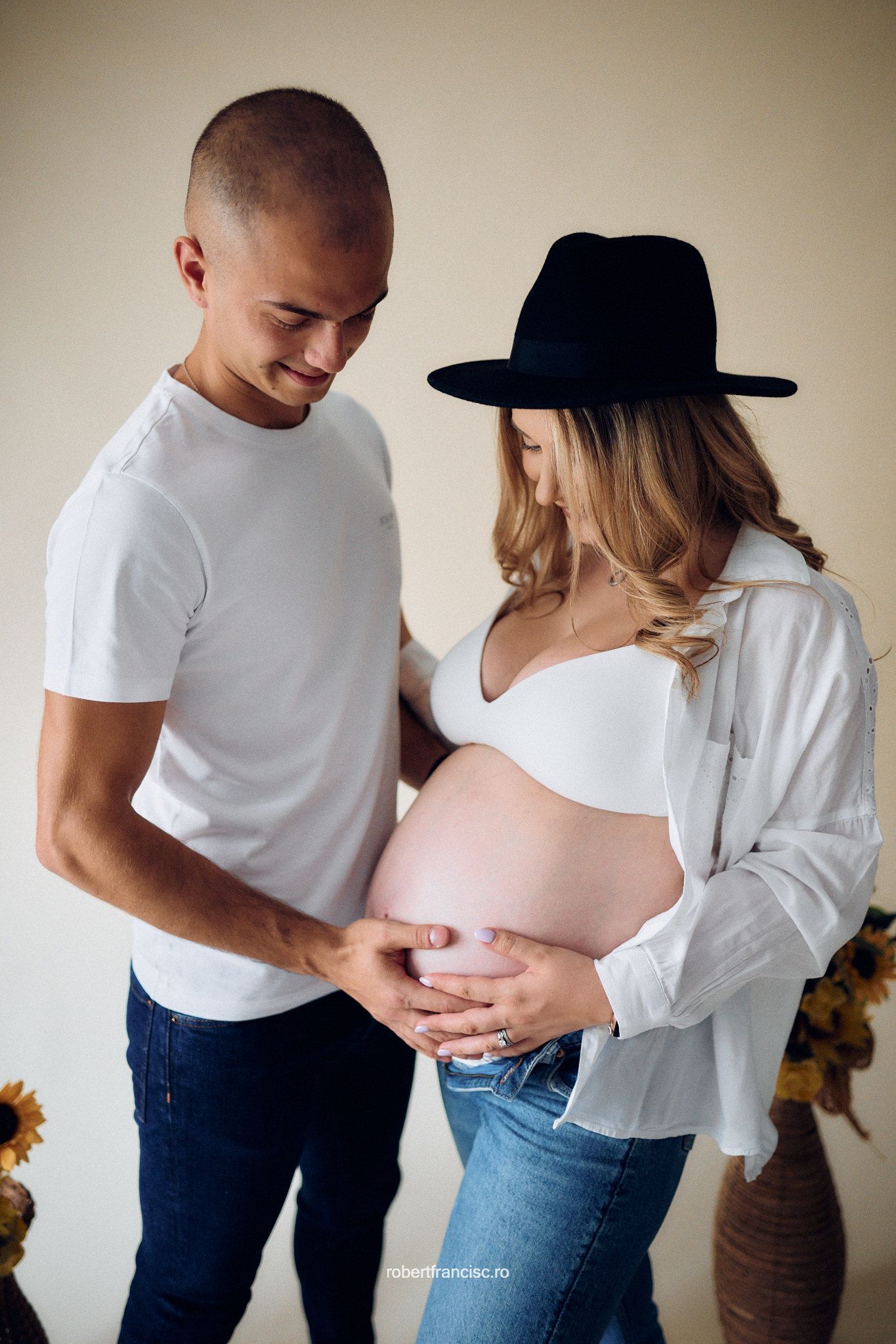 A minimalist maternity photoshoot series showing couple in white tops and jeans against neutral wall with sunflowers.