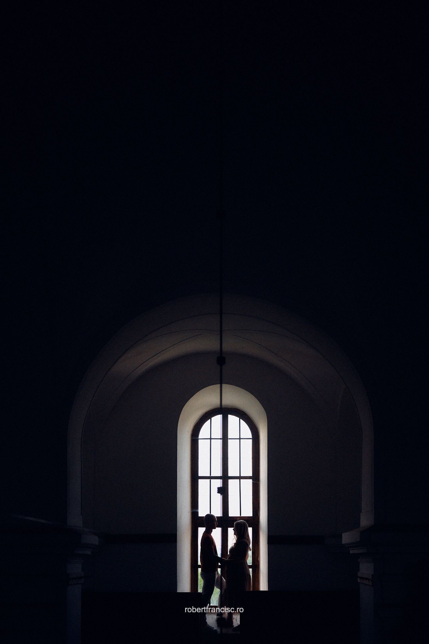 Silhouettes of people stand against a bright arched window in a dark interior space creating dramatic contrast.
