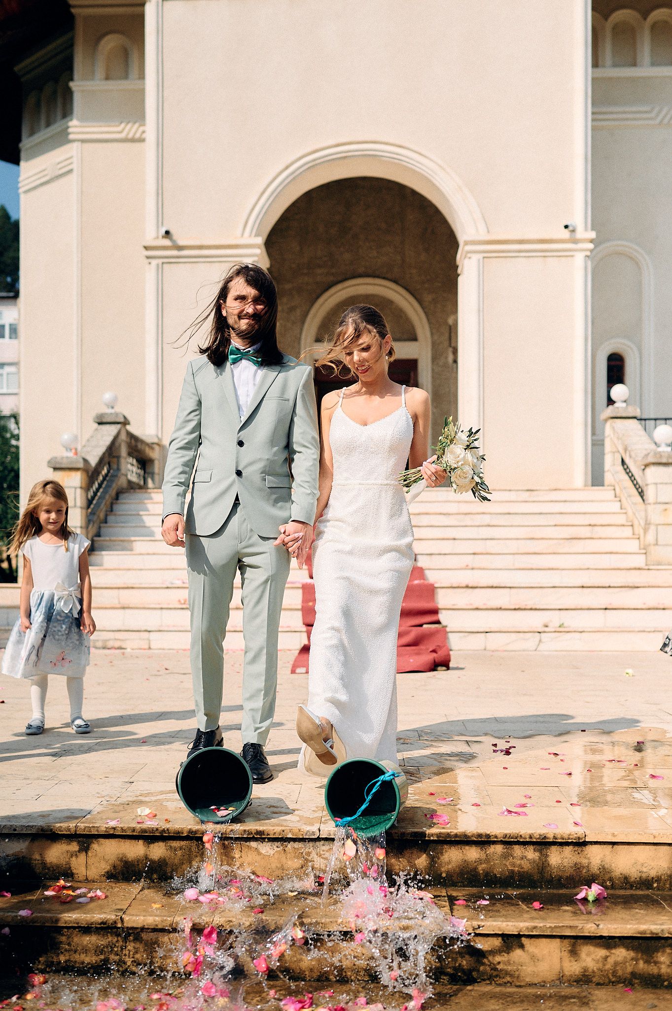 Newlyweds walk down steps by an arched entrance while splashing water from buckets in a playful wedding moment.