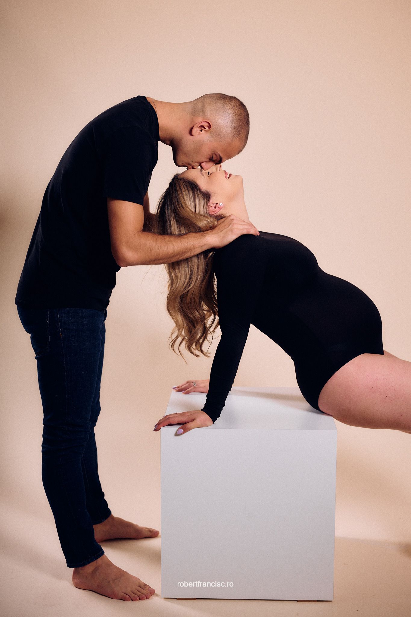 Intimate maternity photo featuring couple in black clothing posing with a white cube against beige background.
