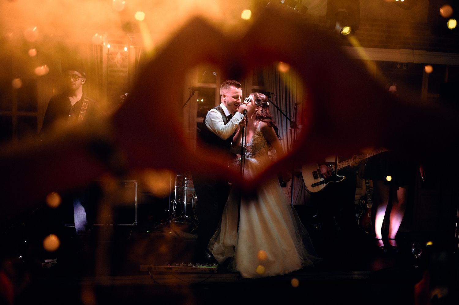 A romantic first dance captured through a heart shape formed by hands with bokeh lights.