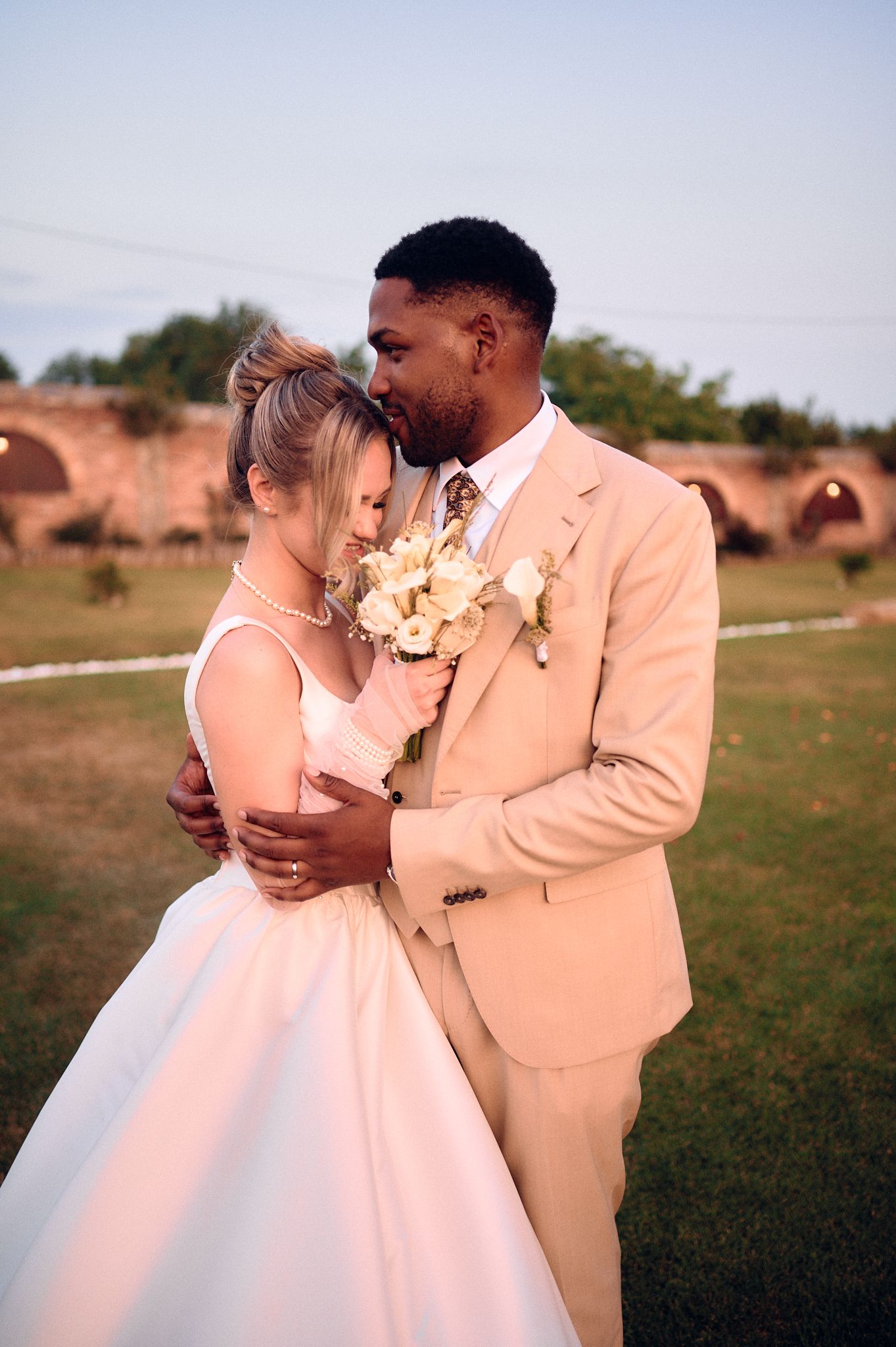 Sunset wedding portrait with couple in peach suit and white dress holding bouquet.