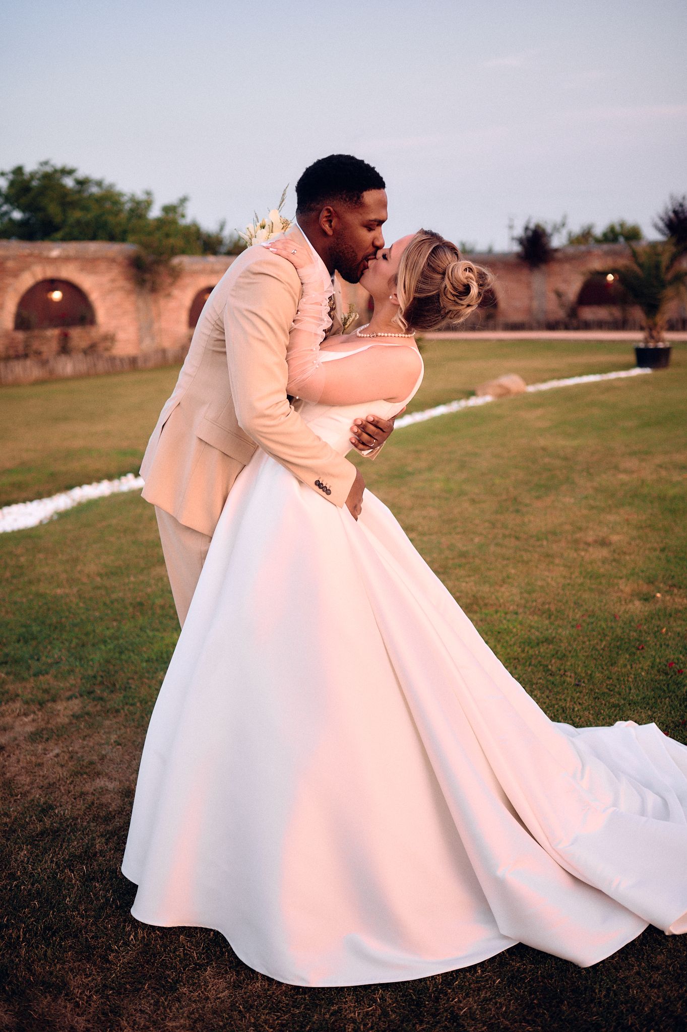Romantic sunset portrait of couple in peach suit and white gown against brick wall backdrop.