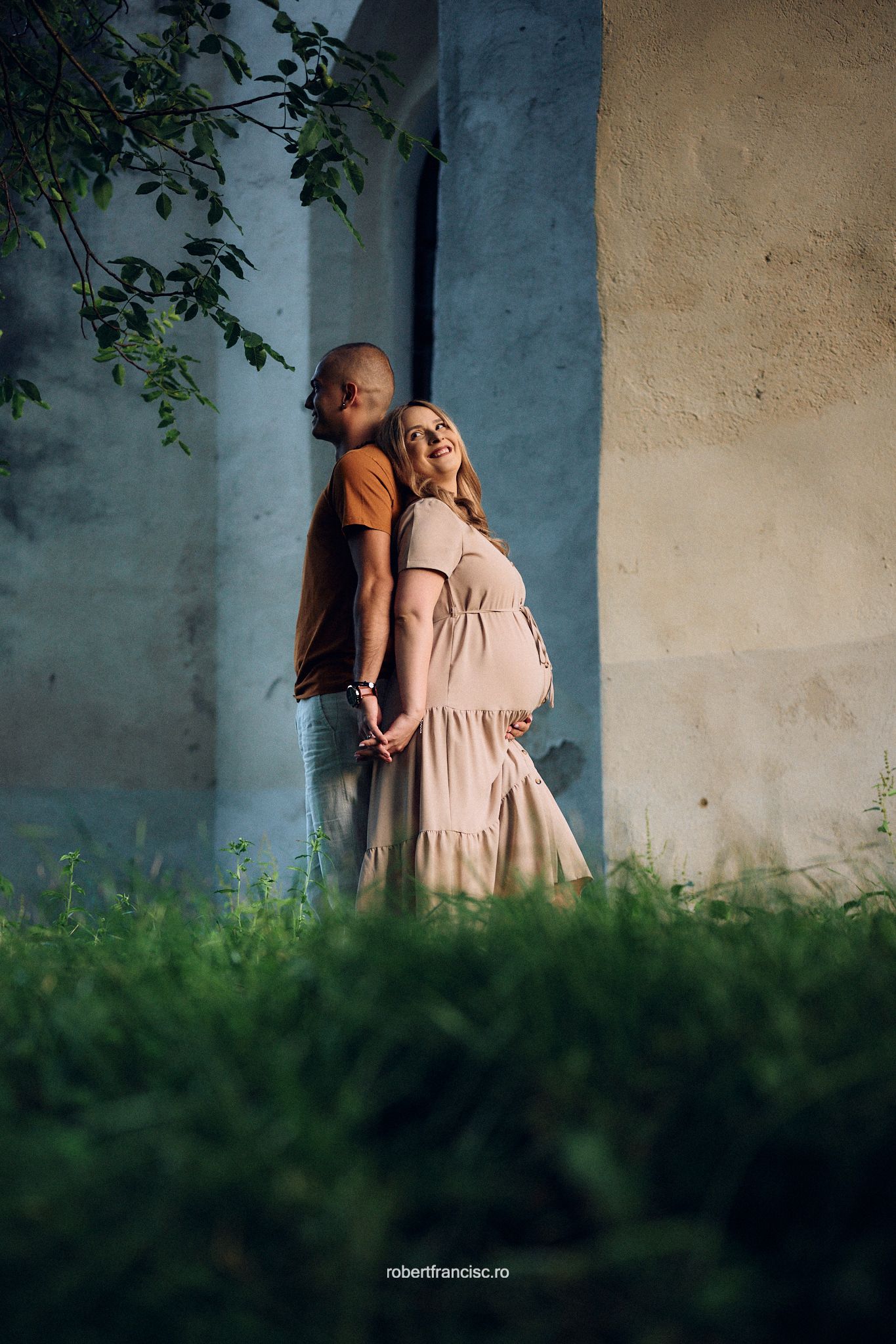 An intimate outdoor portrait shows a couple standing against a textured wall surrounded by greenery.