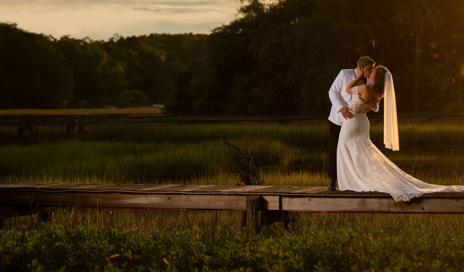 Bride and groom share a romantic kiss on a wooden dock at golden hour with lush green marshlands in the background.