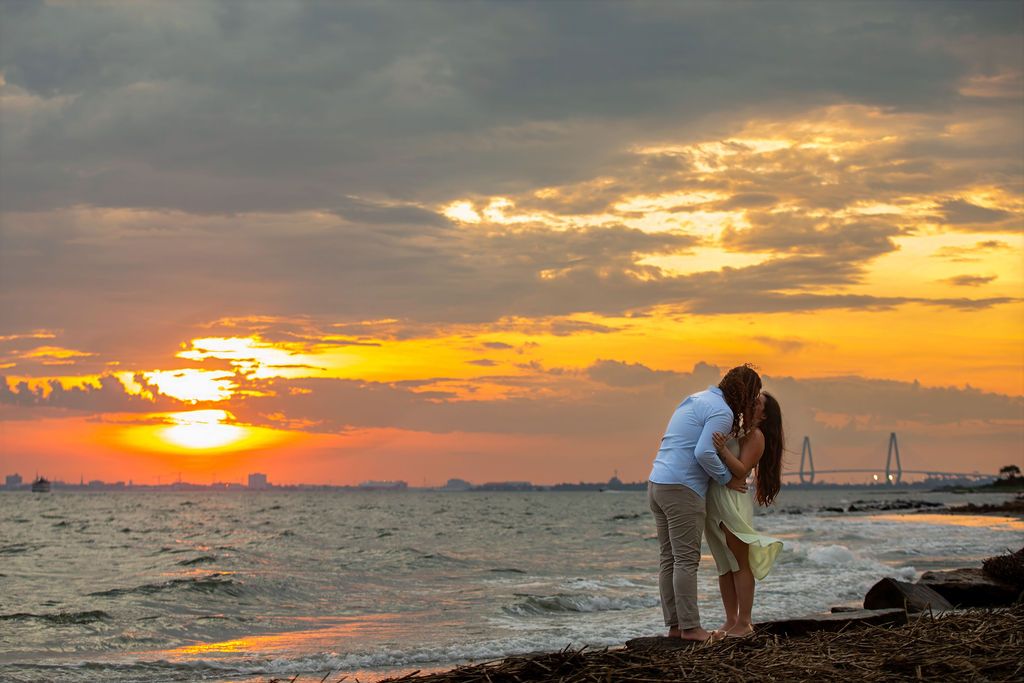 Couple kissing on a rocky beach at sunset with golden sky and distant bridge over calm ocean waters.