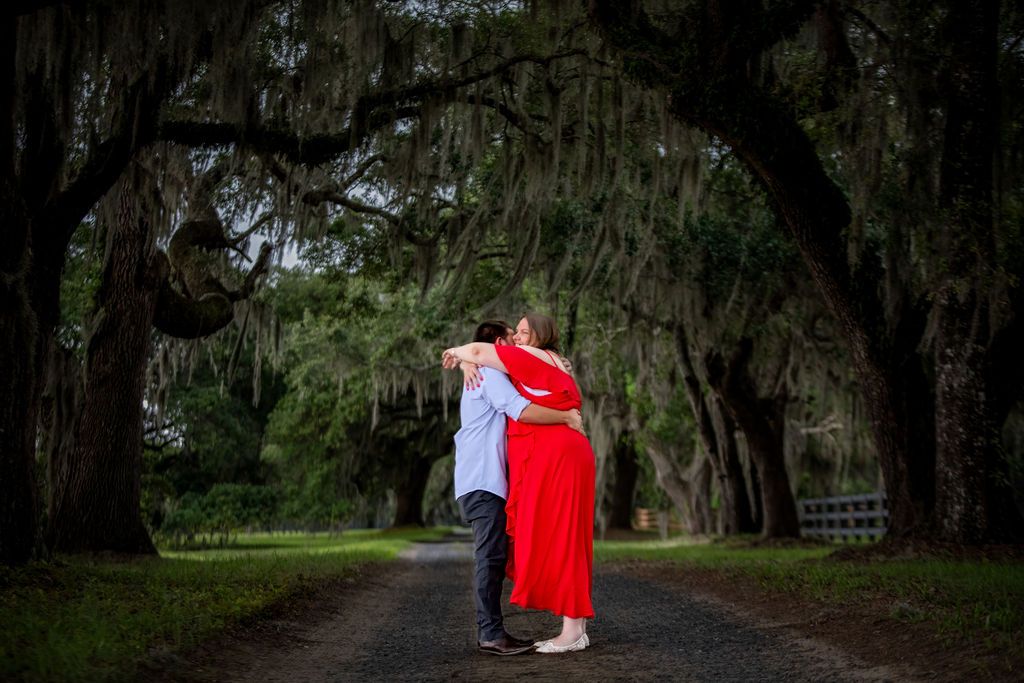 Couple embracing on a moss-draped oak tree lined path, woman in red dress, romantic outdoor engagement photo.