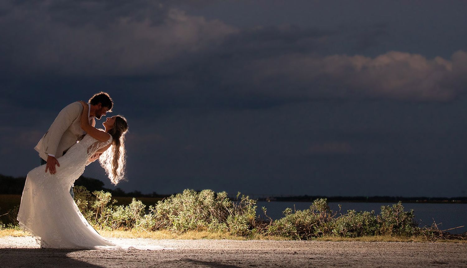 Bride and groom share a romantic dip kiss at dusk by the waterfront under dramatic stormy skies.