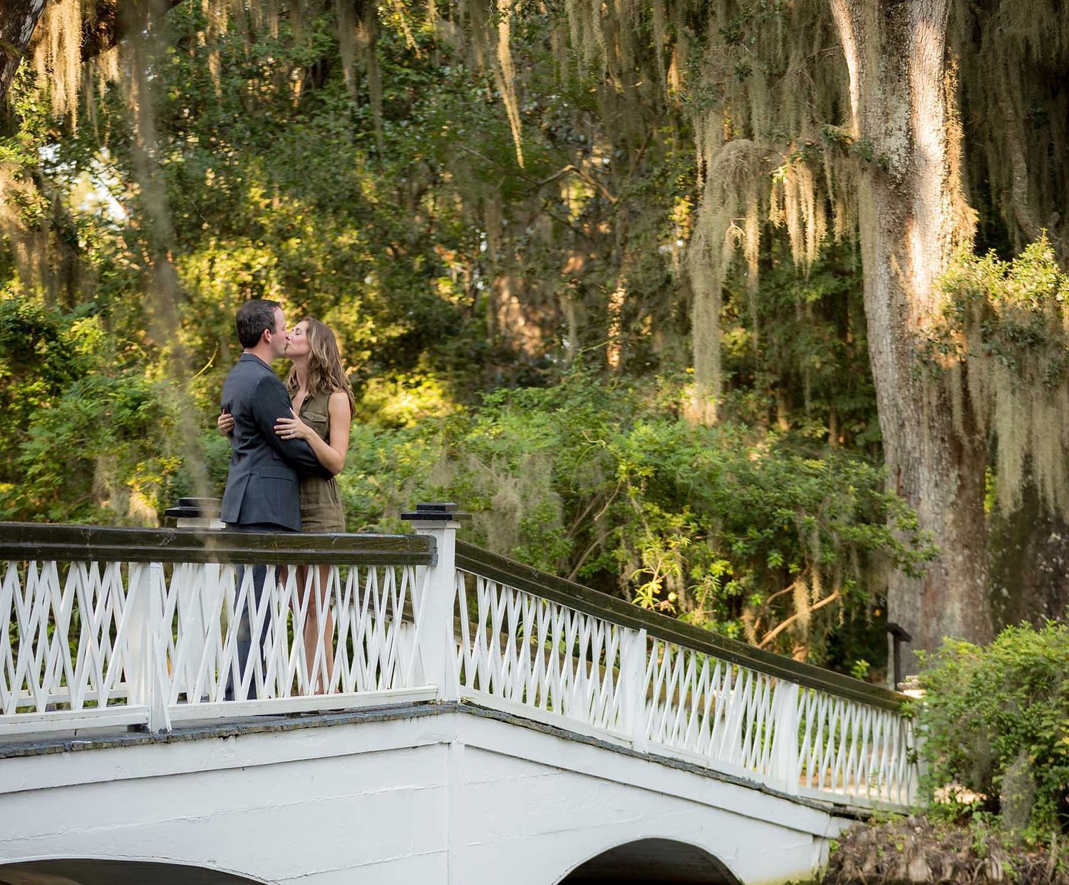 Couple sharing a romantic kiss on a white bridge surrounded by lush trees draped in Spanish moss.