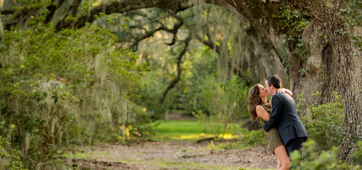 Couple sharing a romantic kiss under a sprawling moss-draped oak tree in a lush green outdoor setting.